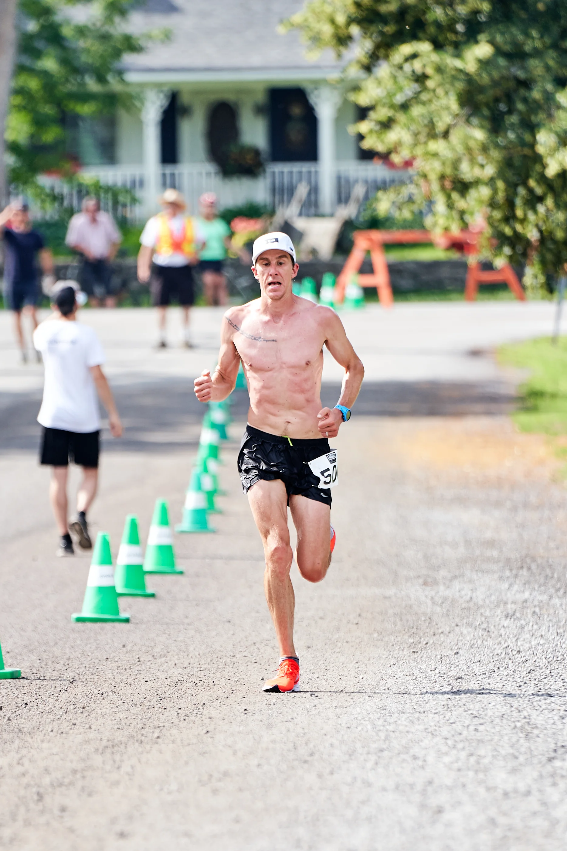 A shirtless man running in a race, wearing black shorts, an orange shoe, a white cap, and a digital watch, with a race number 50 attached to his shorts. The background includes spectators and other runners, with green traffic cones marking the race p
