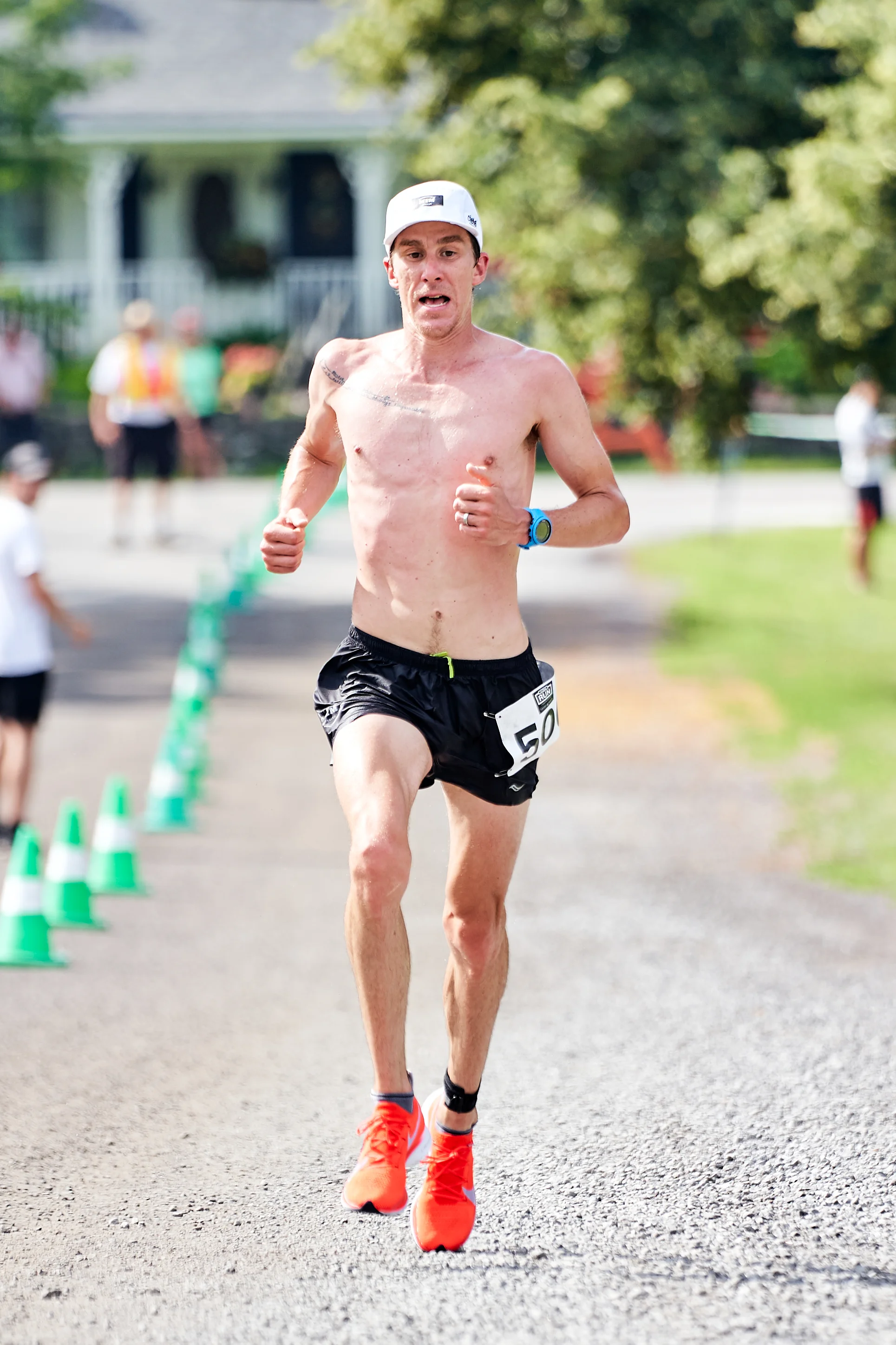 A male marathon runner with a bib number 50, shirtless, wearing a white cap, black shorts, bright orange running shoes, and a blue wristwatch, running on a paved path during a race.