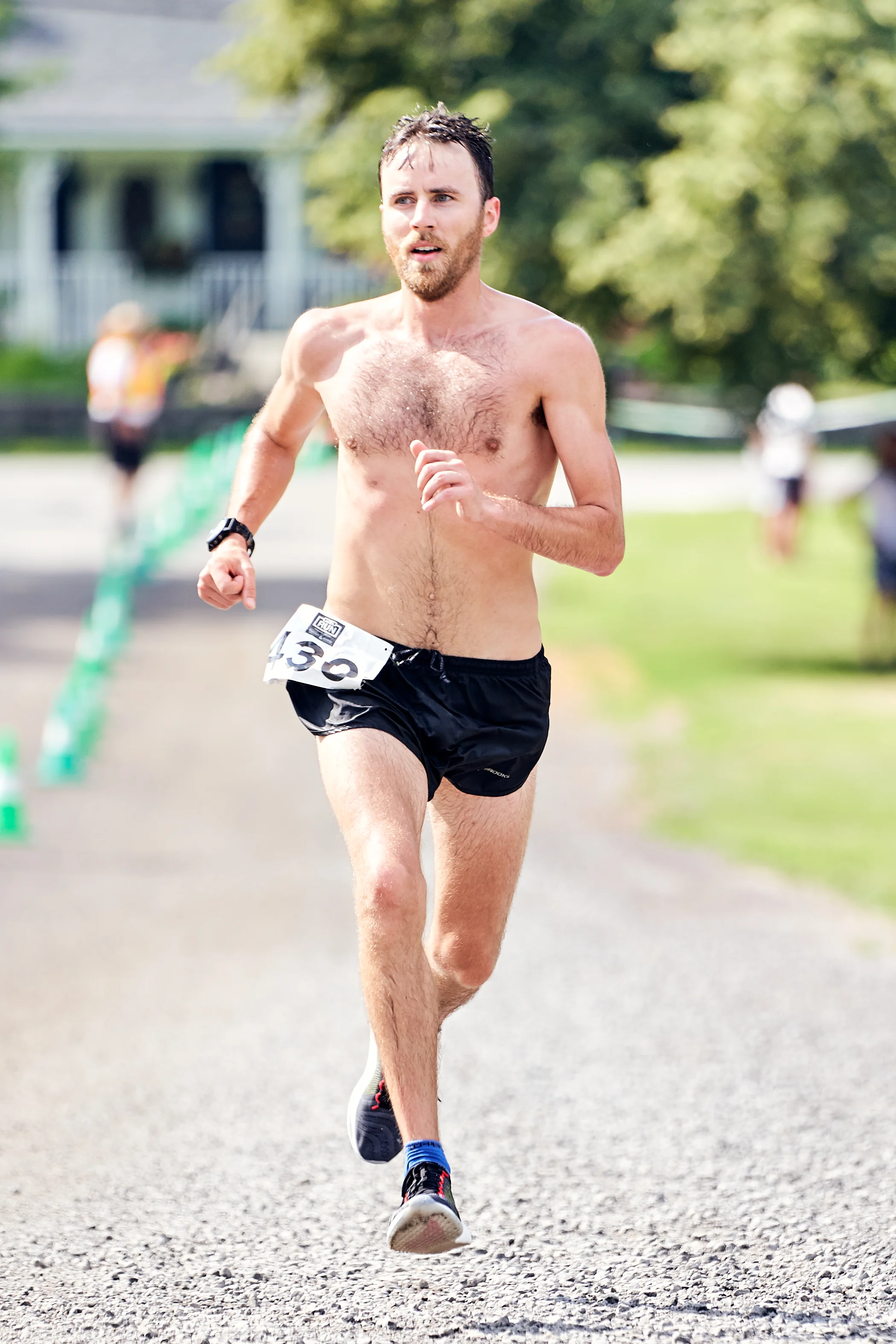 A shirtless man running in a race, wearing black shorts, black running shoes with blue accents, and a race bib with the number 50.
