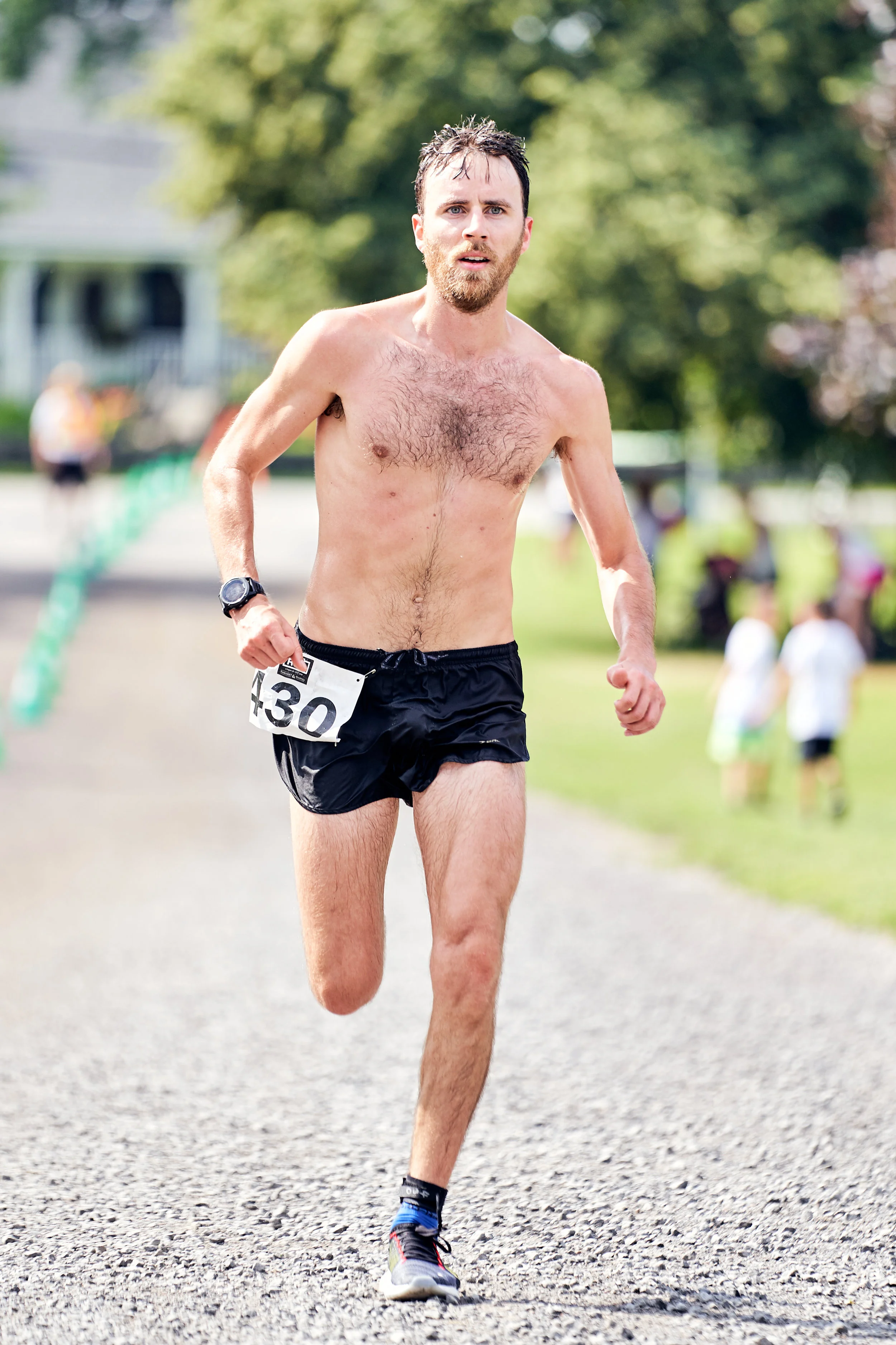 A shirtless man with a beard and race number 430 running outdoors on a gravel path during a race.