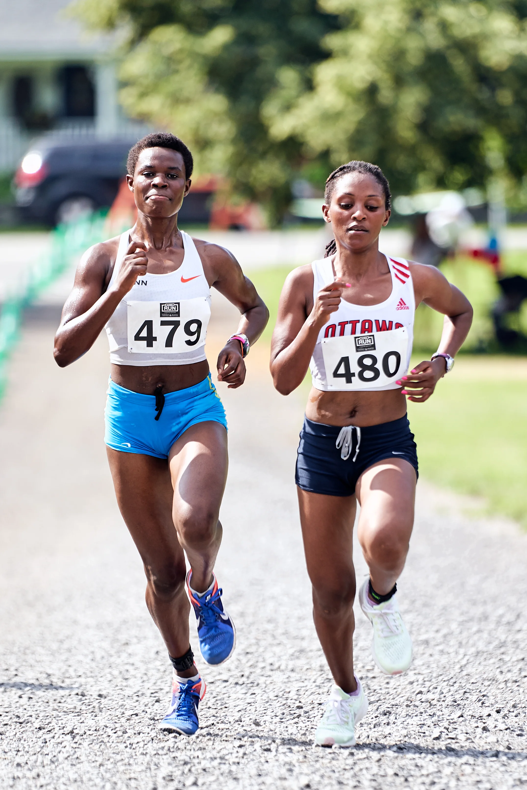 Two female runners competing in a race, running on a gravel path outdoors in warm weather, with trees and vehicles in the background.