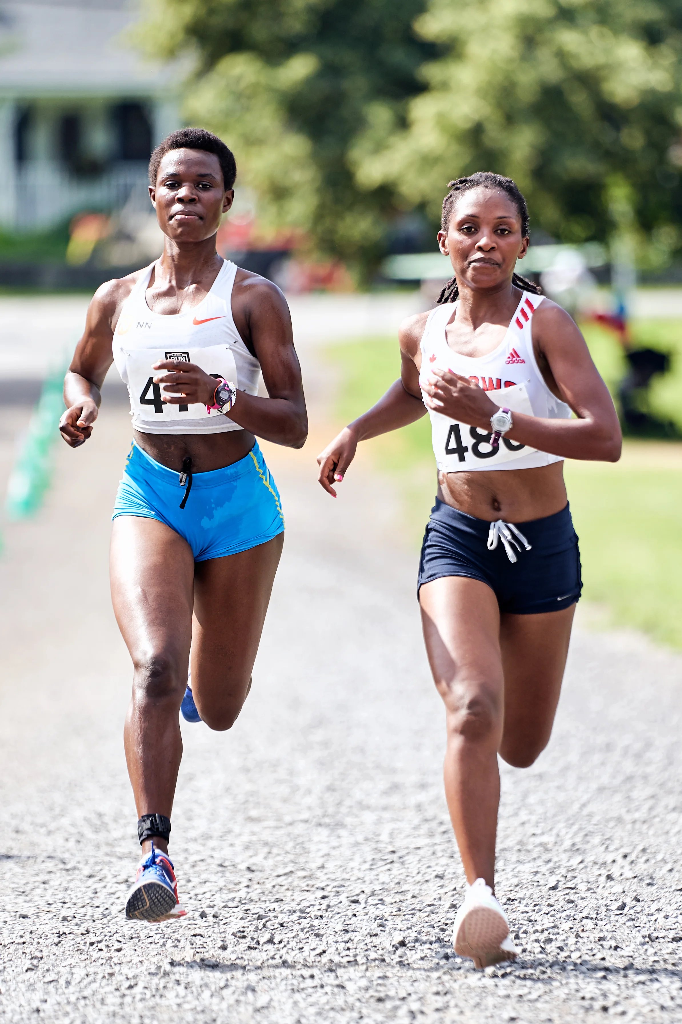 Two female athletes running in a race outdoors on a gravel track, wearing athletic shorts and tops with race numbers 43 and 48, with trees and a house in the background.