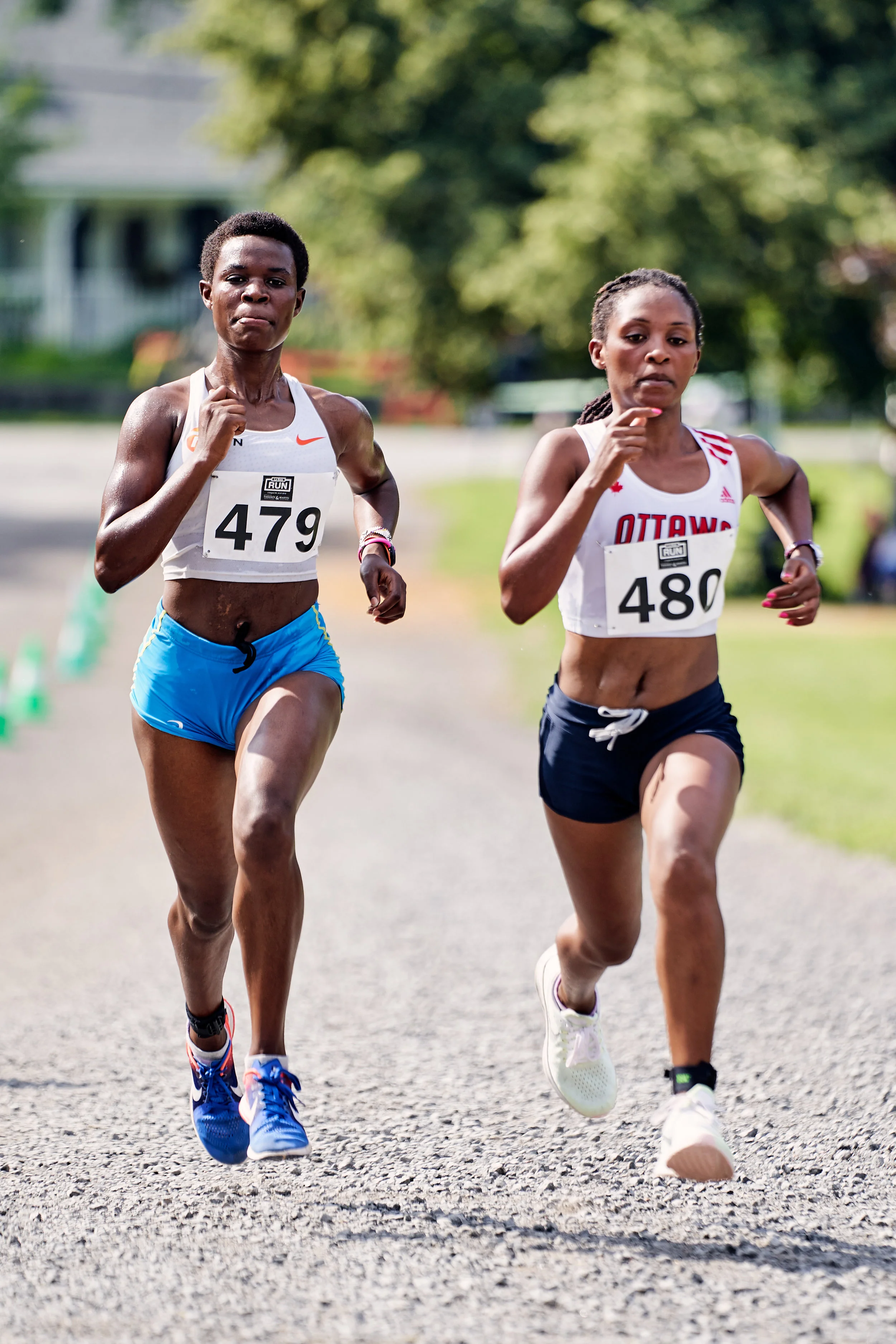 Two female runners competing in a race outdoors, with trees and a house in the background.