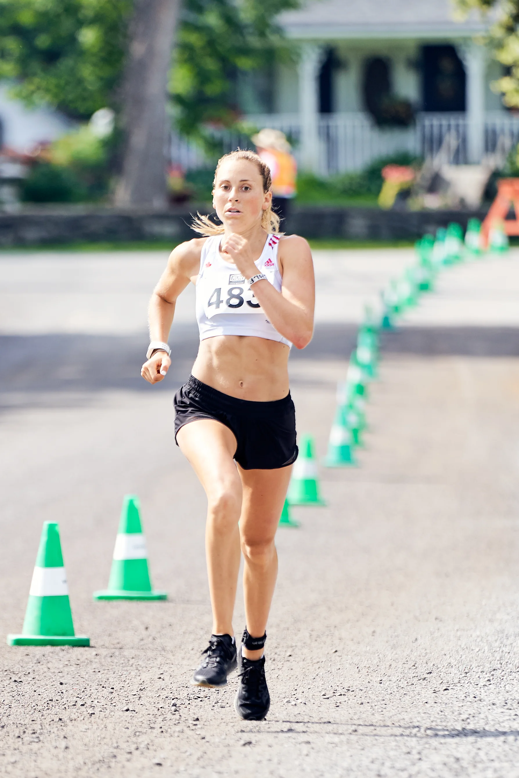Female runner in a race, wearing bib number 483, running on an outdoor track lined with green cones and orange barriers, with trees and houses in the background.