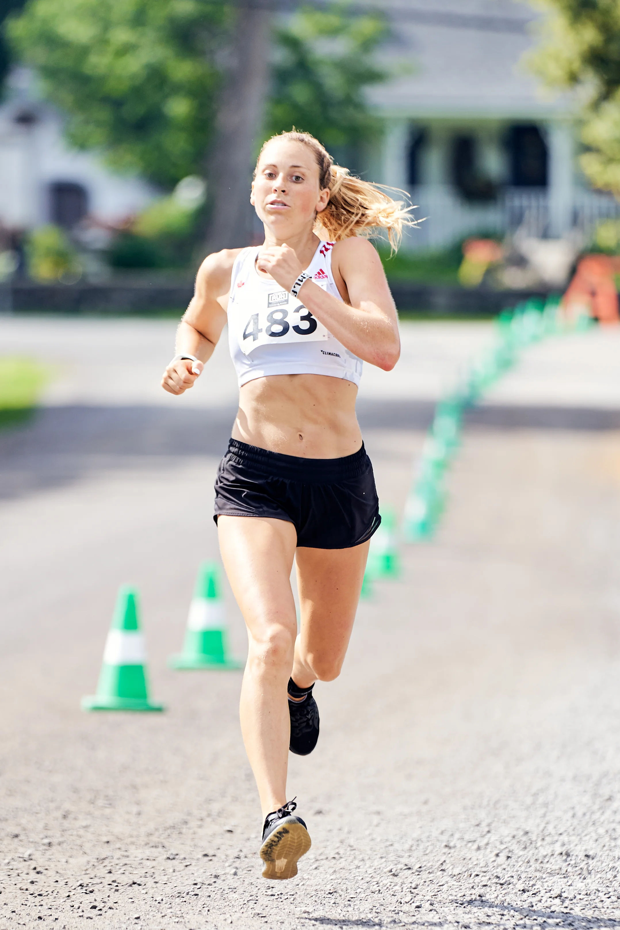 A female runner in a race, wearing a white sports bra with the number 483 and black shorts, running on a paved road with green and white cones.