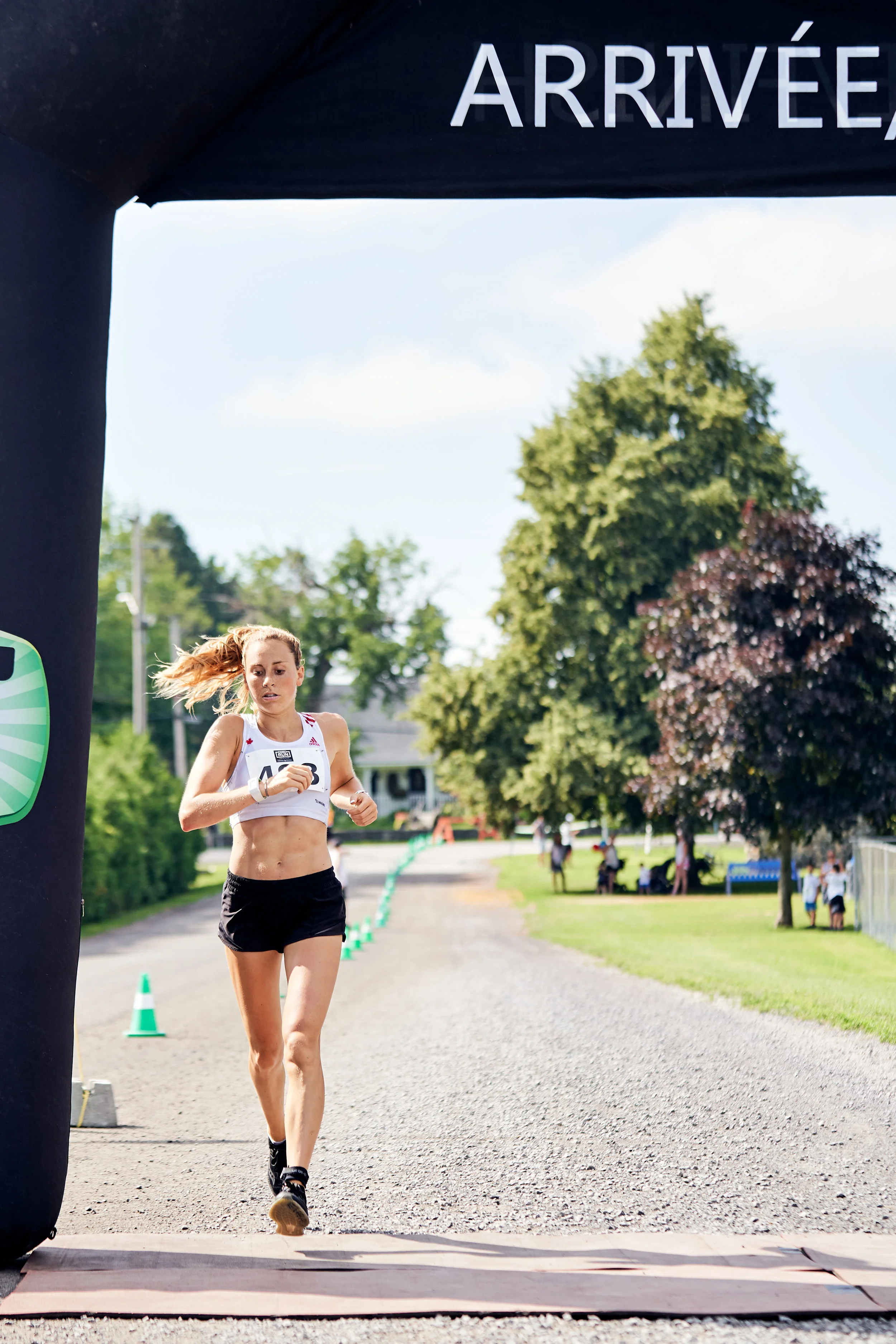 A female runner crosses the finish line at an outdoor race, with trees and spectators in the background, under a black inflatable archway with the word 'ARRIVÉE' on it.