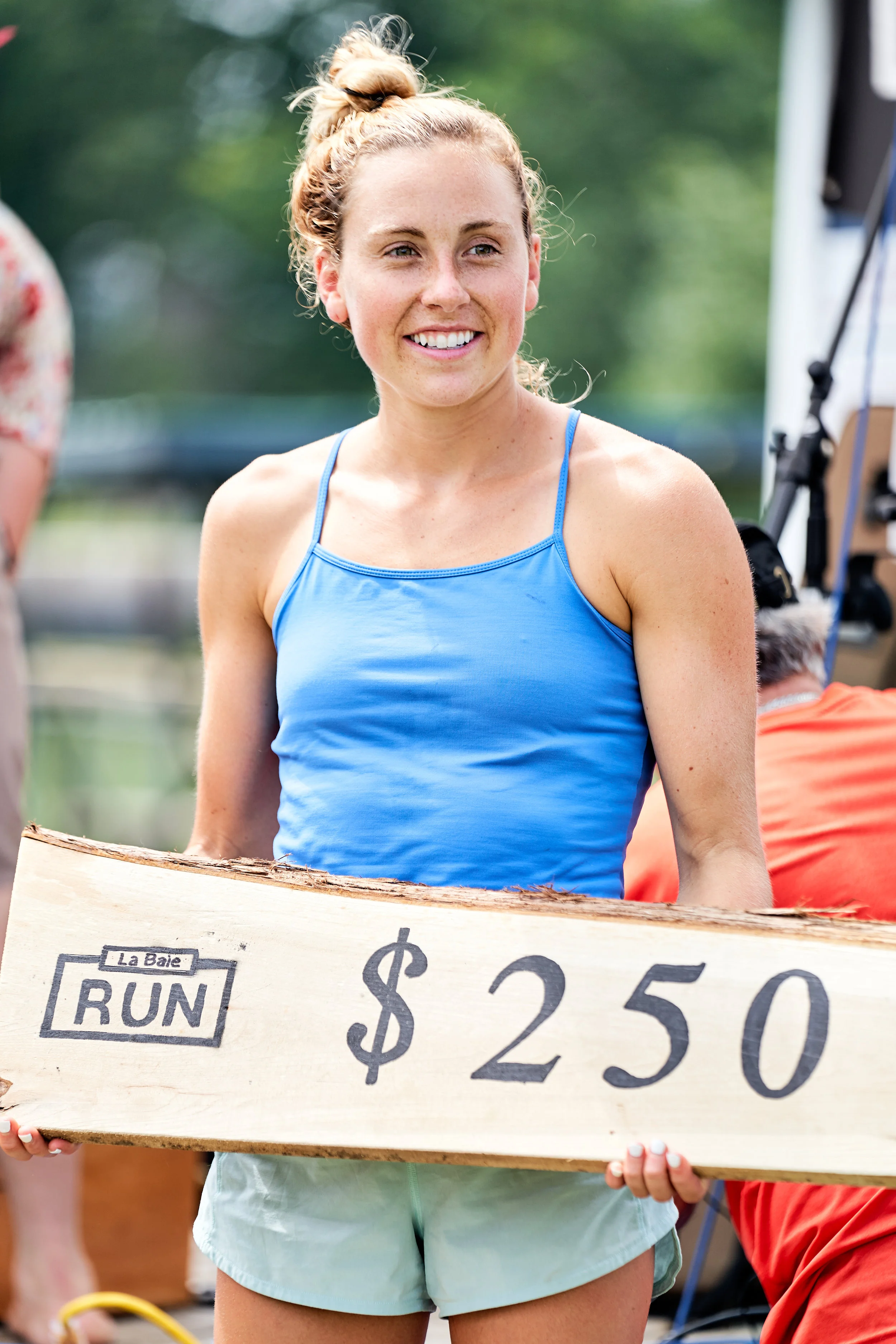 A woman smiling and holding a wooden sign that reads '$2.50' for a charity run at La Baie.