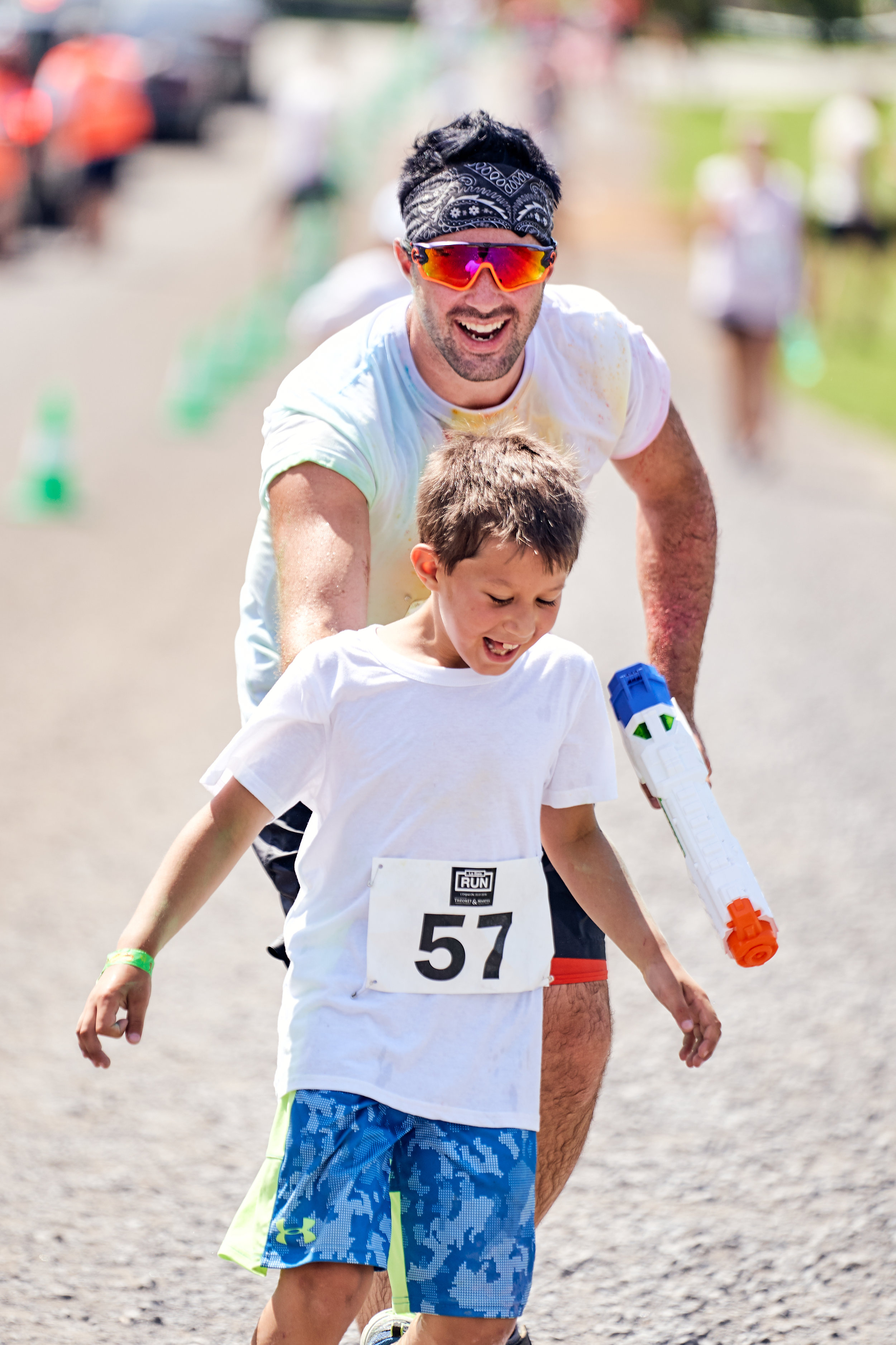 A man and a young boy participating in a relay race, with the boy holding a toy water gun, on an outdoor running trail during a sunny day.