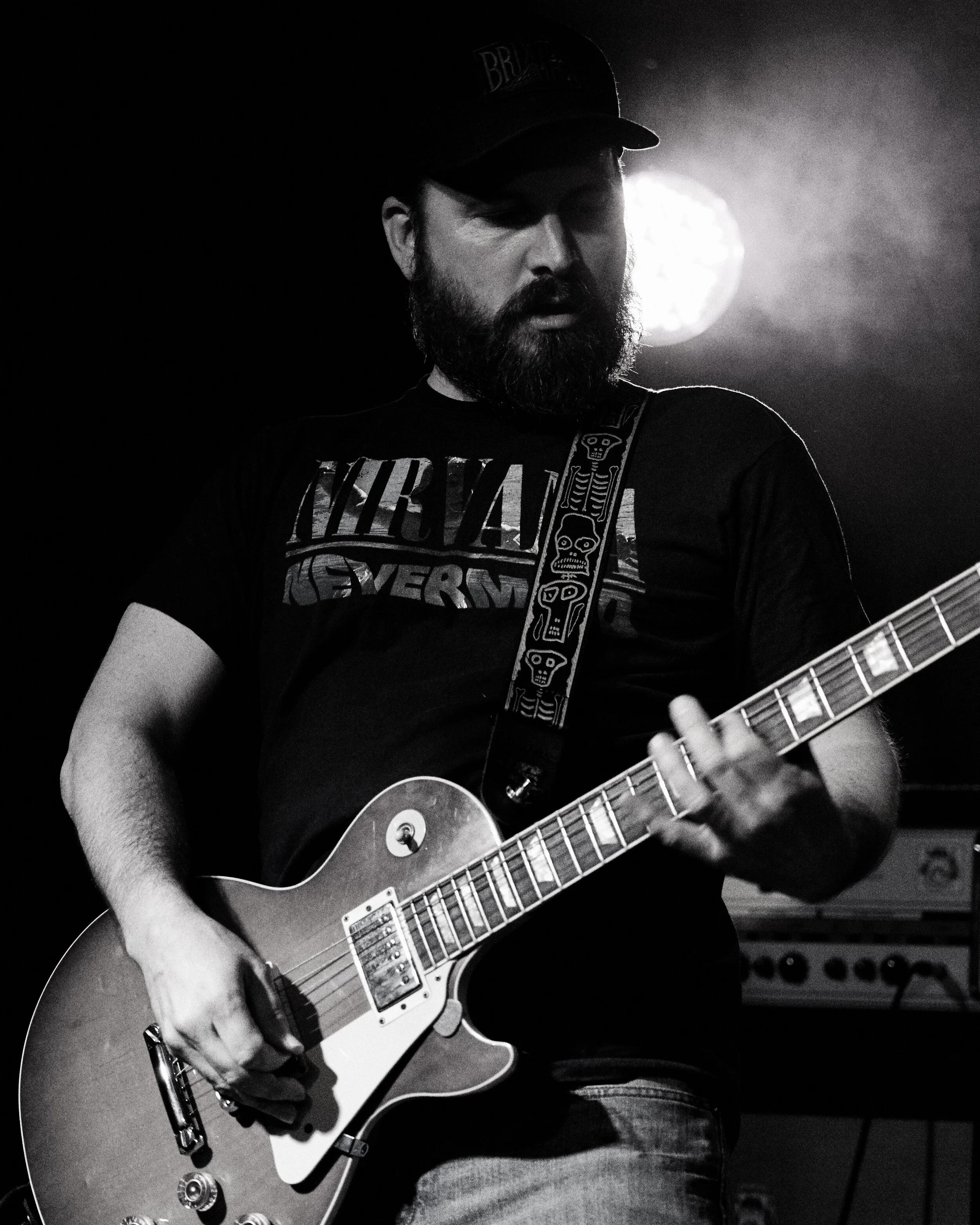 A bearded man wearing a baseball cap and Nirvana t-shirt playing an electric guitar on stage with a bright light behind him.