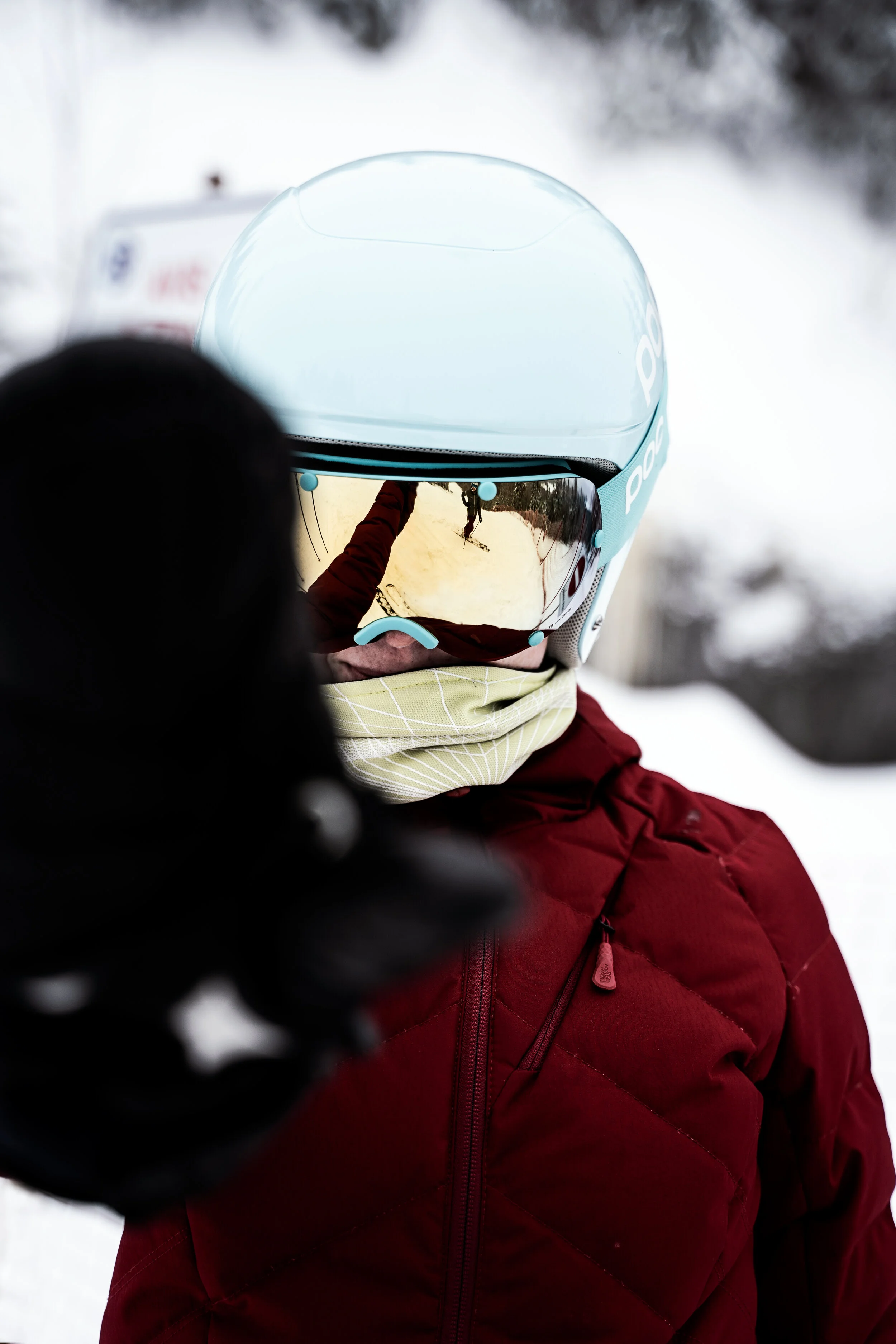 Person wearing ski gear, including a teal helmet, reflective ski goggles, and a red jacket, in a snowy outdoor setting.