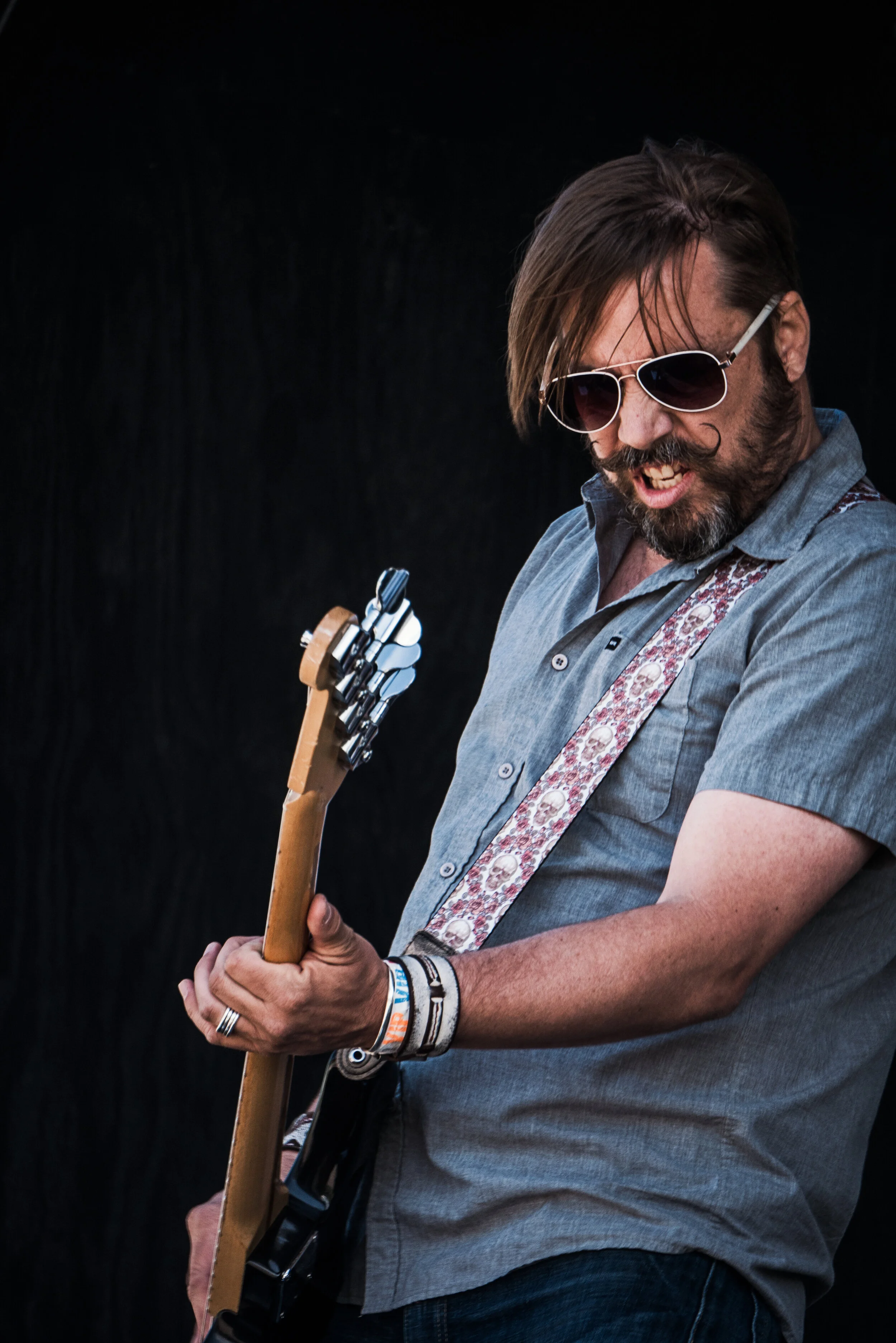 A man wearing sunglasses and a gray short-sleeve shirt plays an electric guitar, with a black background.