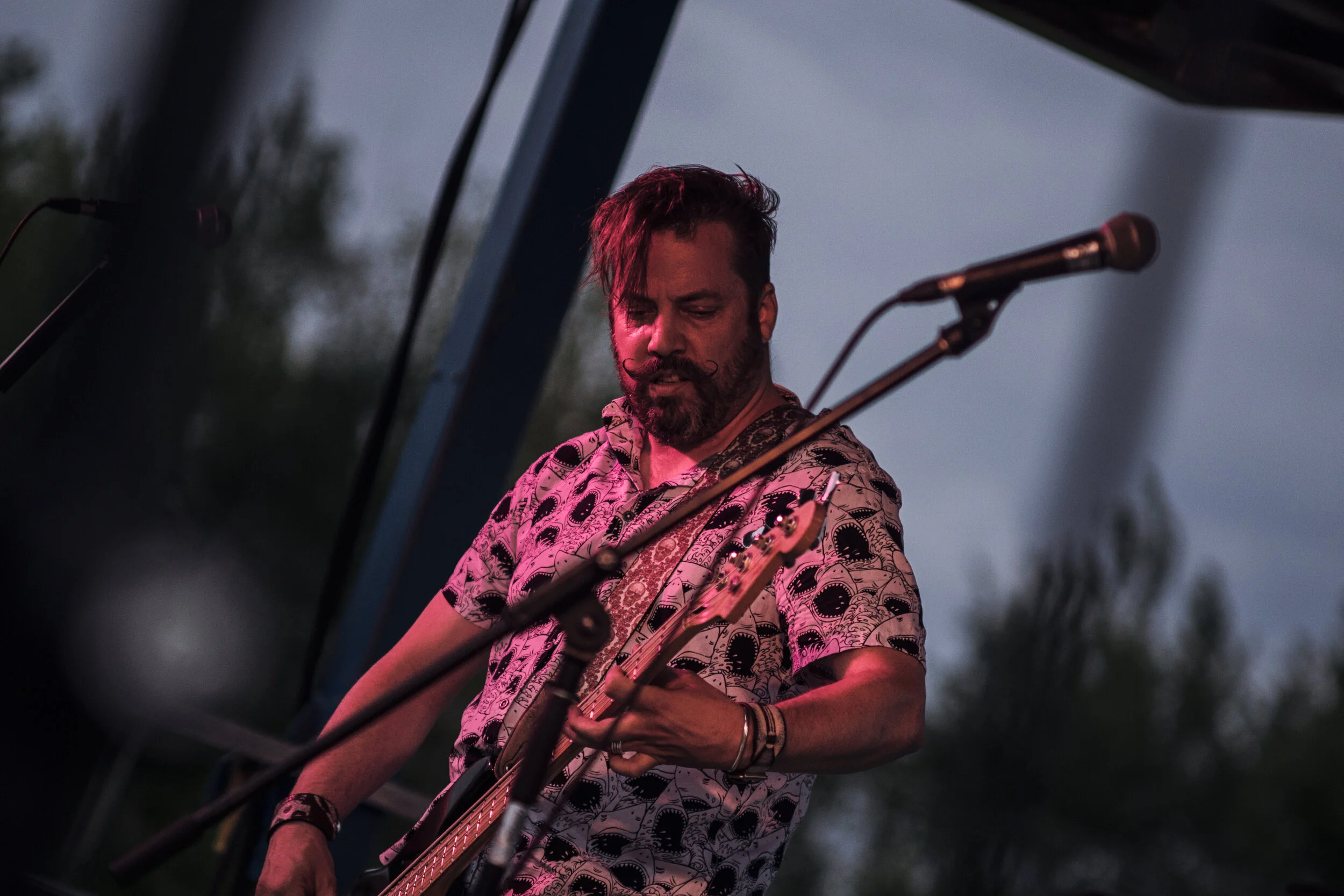 A male musician with dark hair and a beard, wearing a patterned shirt, playing a guitar on an outdoor stage during dusk.