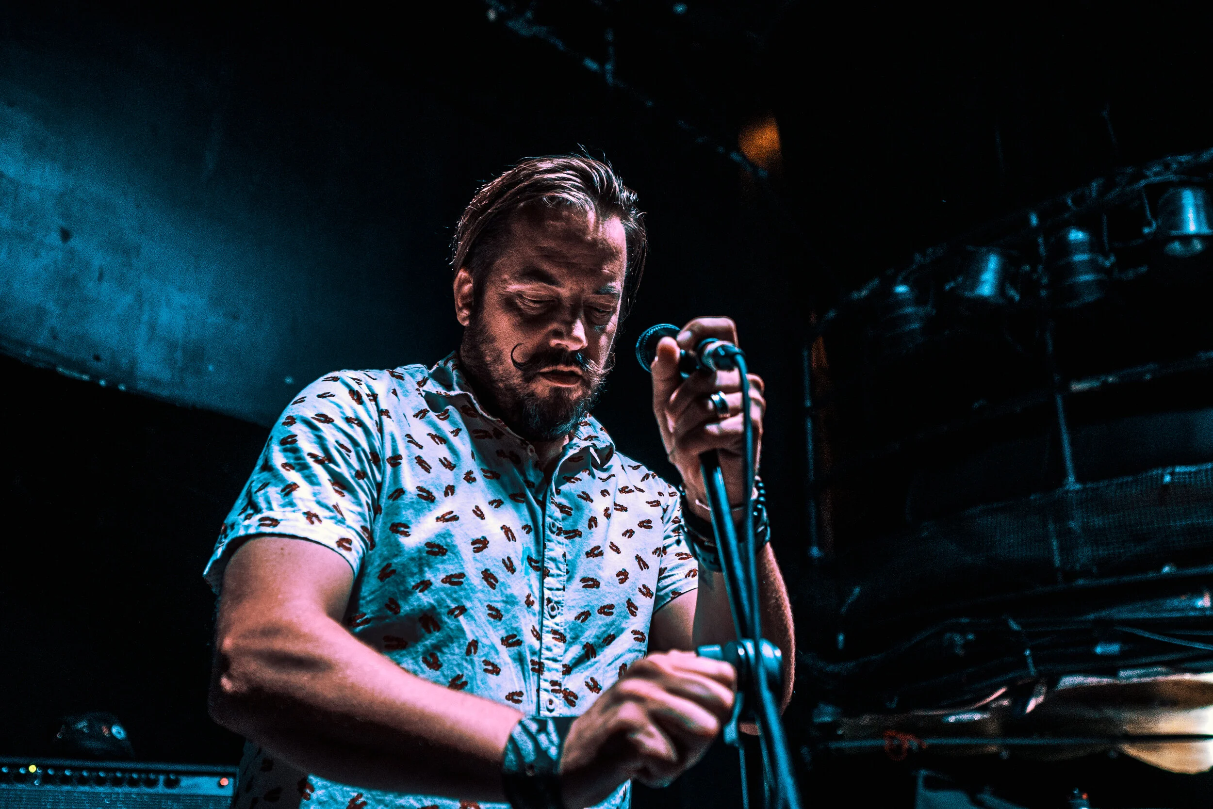 A man with a beard and mustache, wearing a patterned short-sleeve shirt, holds a microphone on stage with dark background lighting.