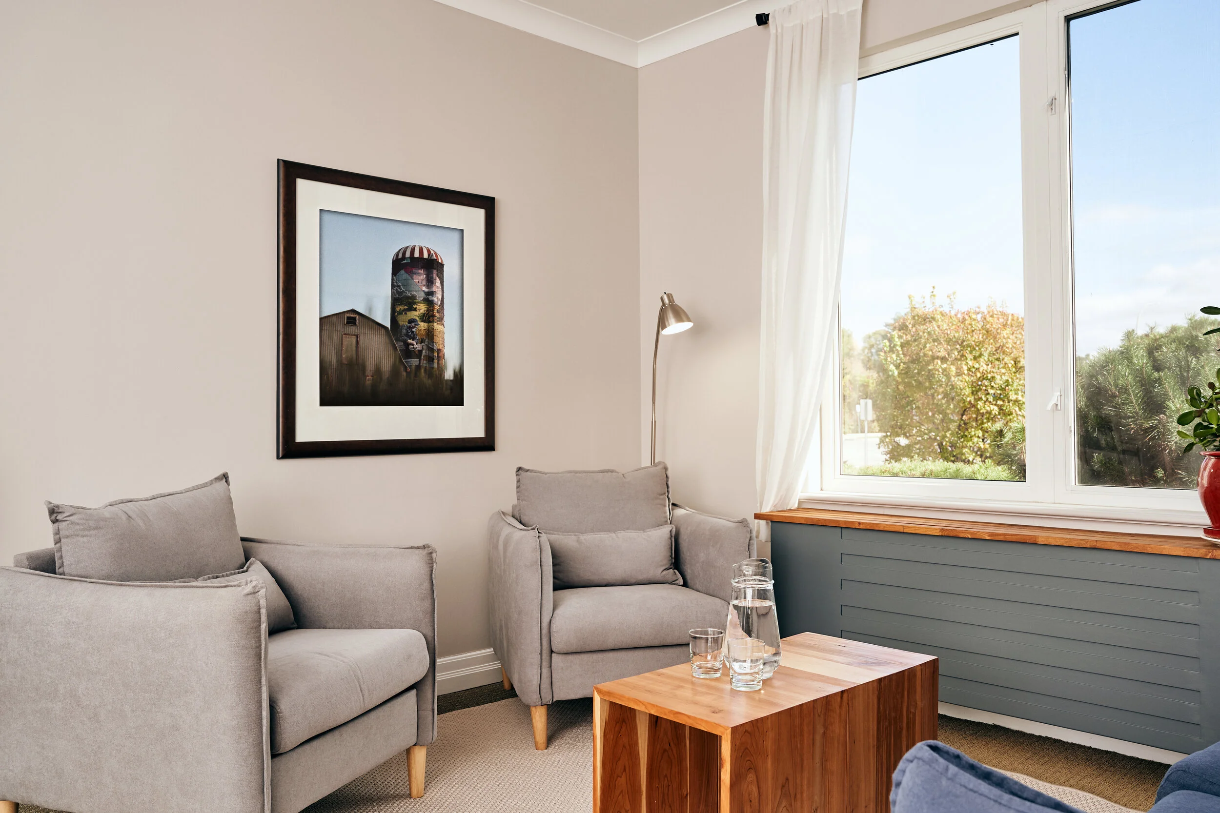 Living room with gray armchair and sofa, wooden side table with water pitchers, large window with white curtains, framed farm photograph on wall, floor lamp, potted plant