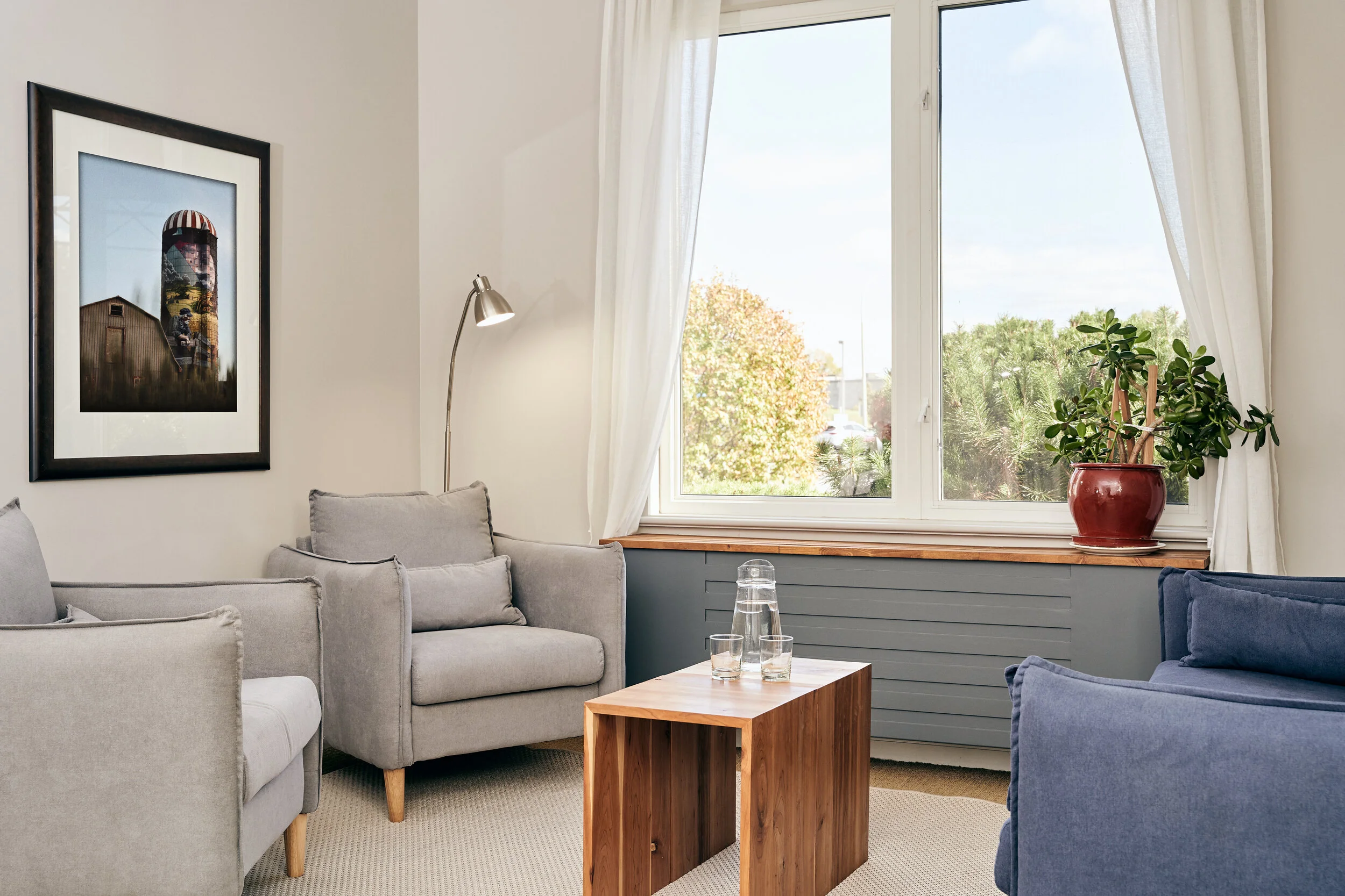 A cozy living room with gray and blue sofas, a wooden coffee table with a glass pitcher and glasses, a large window with white curtains, a potted plant on the windowsill, a framed picture on the wall, and a floor lamp.