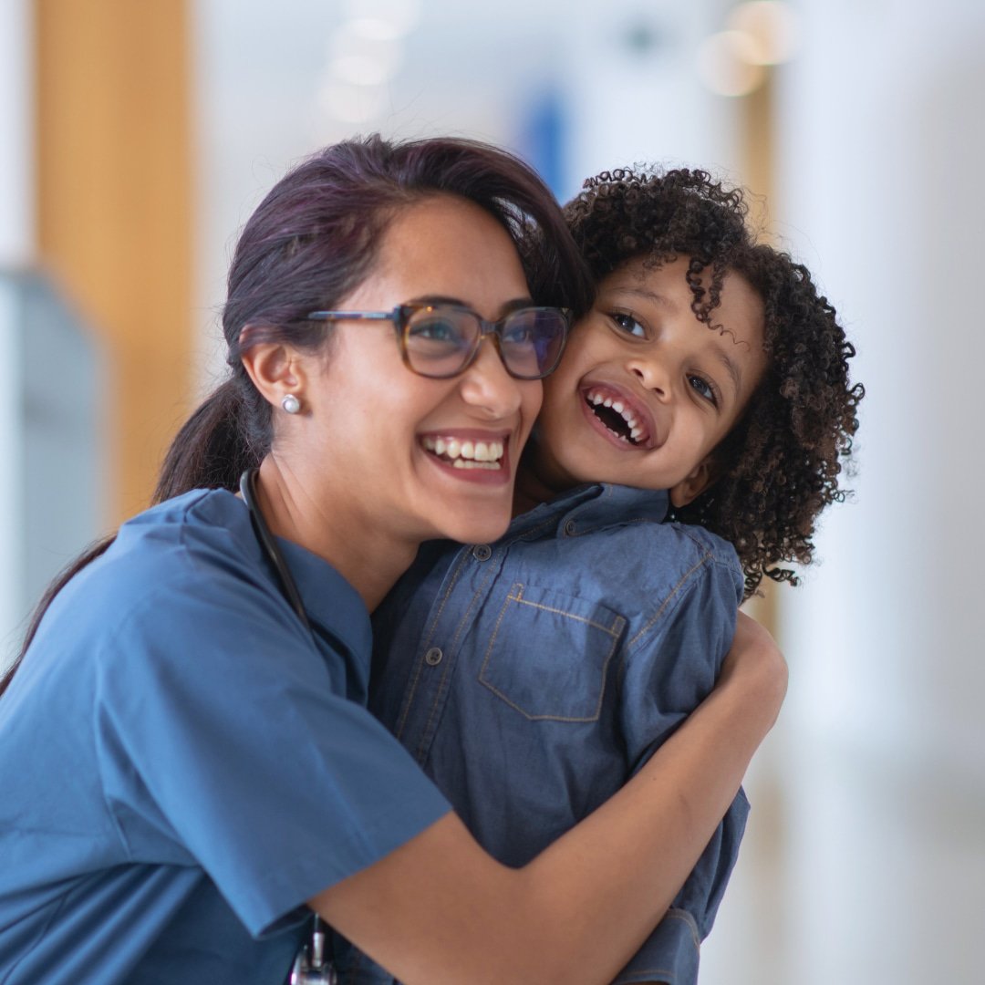 A healthcare worker wearing glasses and blue scrubs hugging a smiling young child with curly hair in a medical facility corridor.