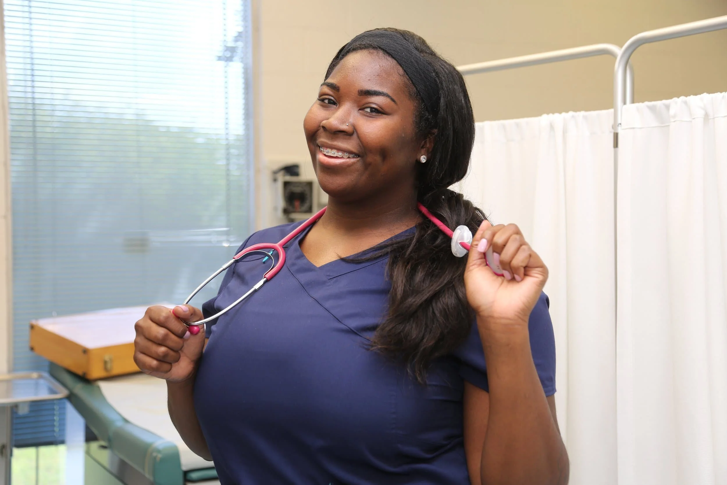 A smiling nurse in navy scrubs holding a stethoscope with pink tubing over her shoulder in a healthcare setting.