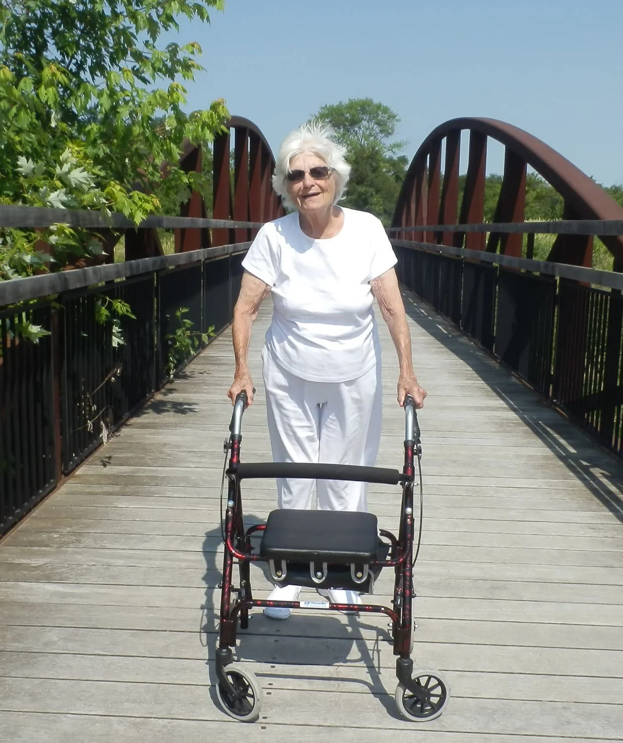 An elderly woman with white hair, wearing sunglasses, a white shirt, and white pants, stands on a wooden bridge with a walker in front of her on a sunny day. Green trees and a clear sky are in the background.
