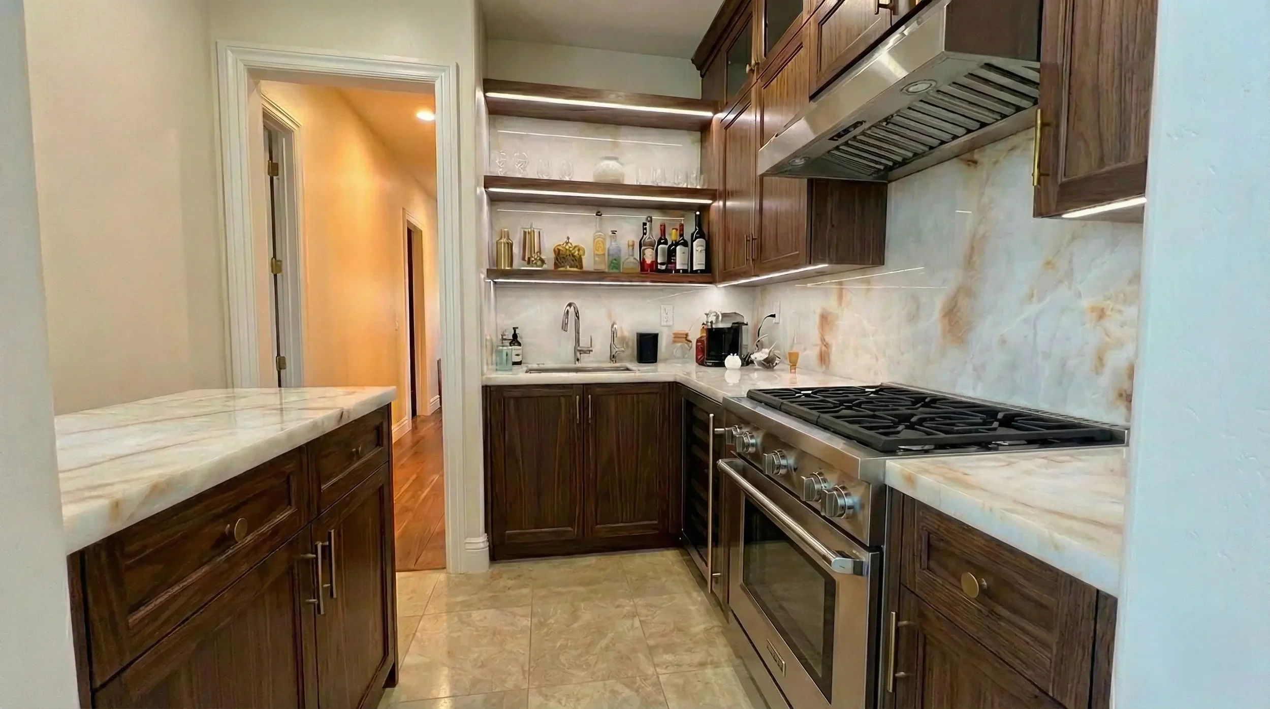 Modern galley kitchen with dark wood cabinets, marble-style countertops and backsplash, stainless steel gas range and hood, open shelves displaying liquor bottles and glassware, and a compact sink area with a coffee station.