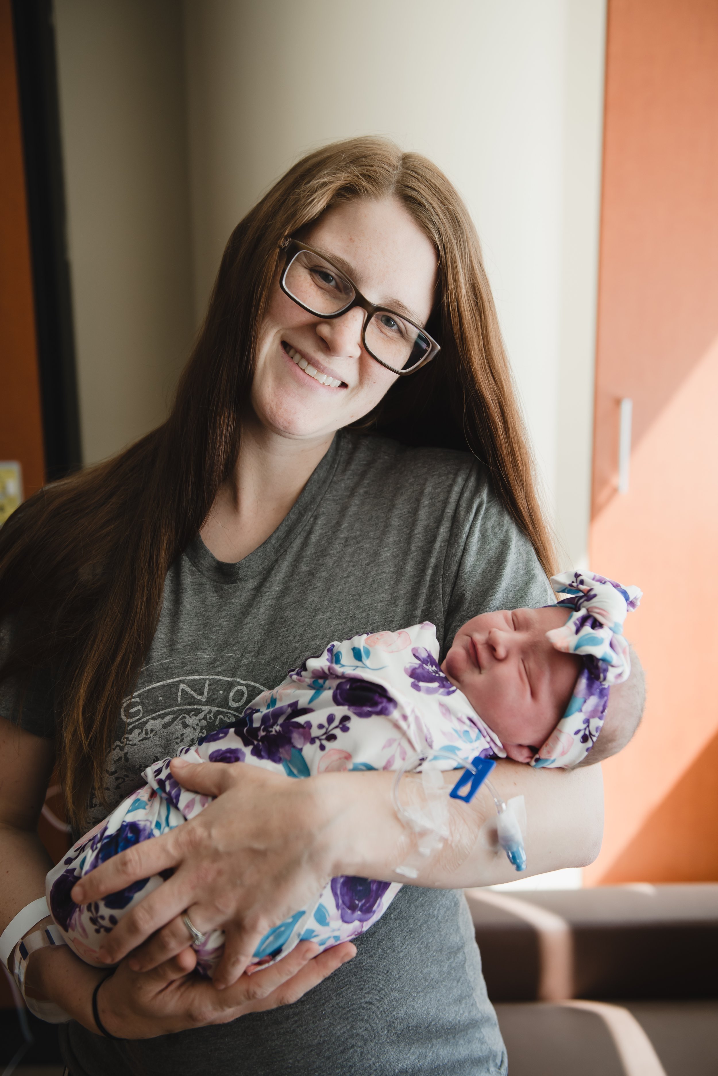 A smiling woman with long red hair, glasses, and a gray t-shirt holding a newborn baby wrapped in a floral blanket in a hospital room.