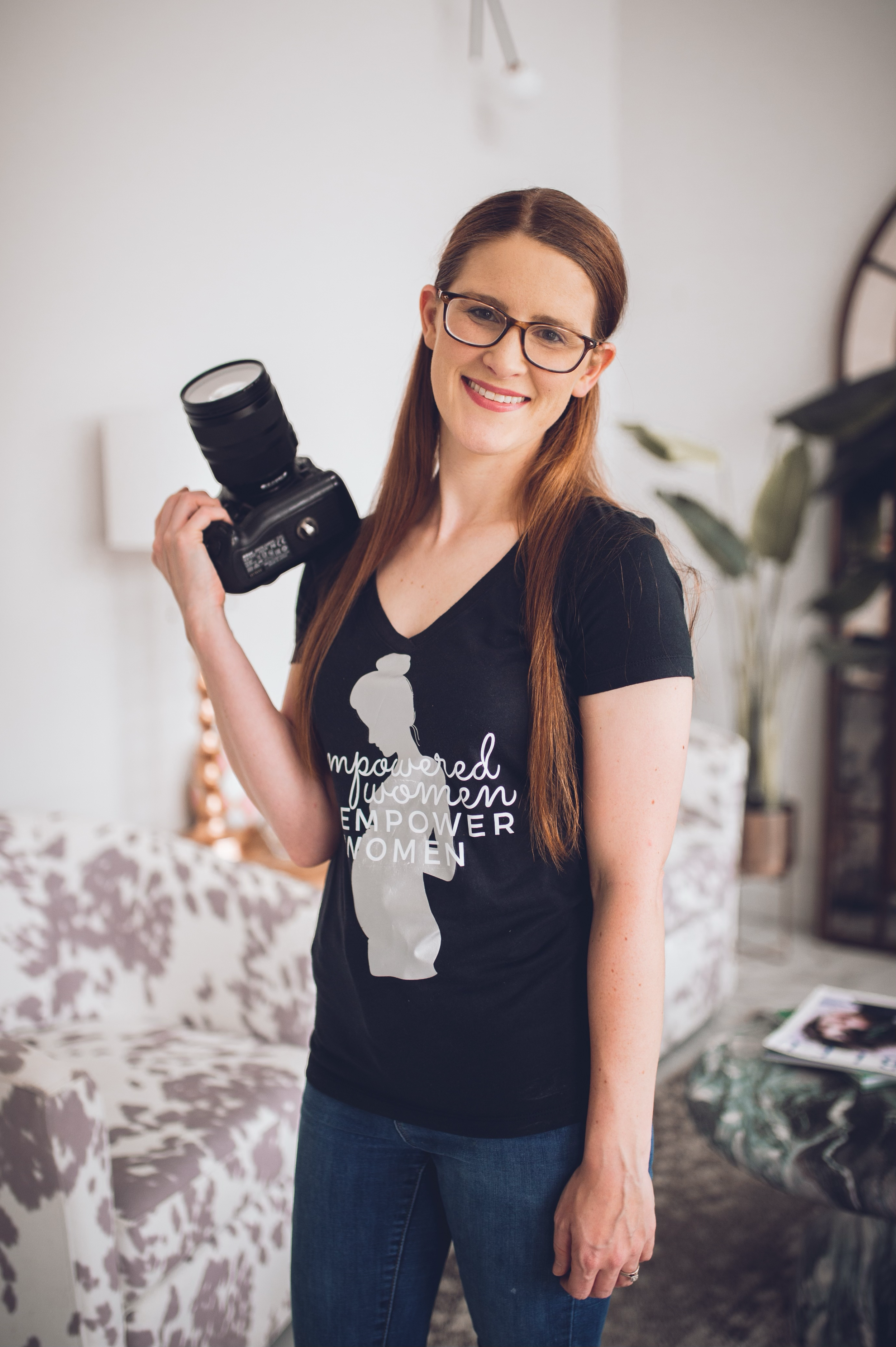 A woman with long red hair, glasses, and a smile holds a camera over her shoulder, standing in a well-lit living room.