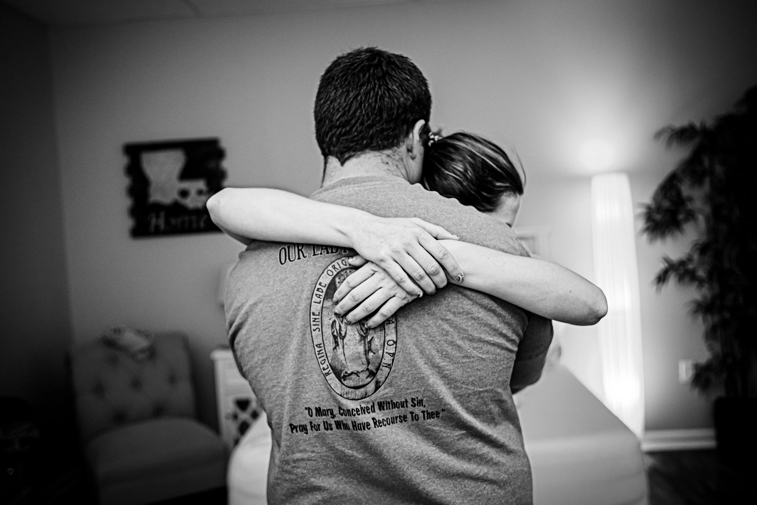 A man and woman hug in a softly lit room, with the woman’s arms around the man's neck and the man’s back to the camera, revealing a shirt with a circular emblem and text.