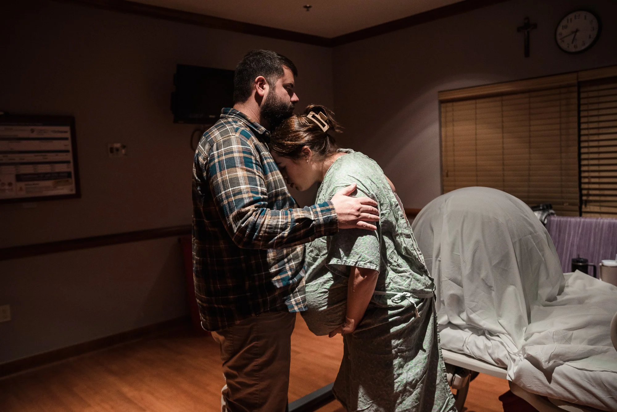 A man and a woman in a hospital room, holding each other in a comforting embrace, with the woman appearing distressed.