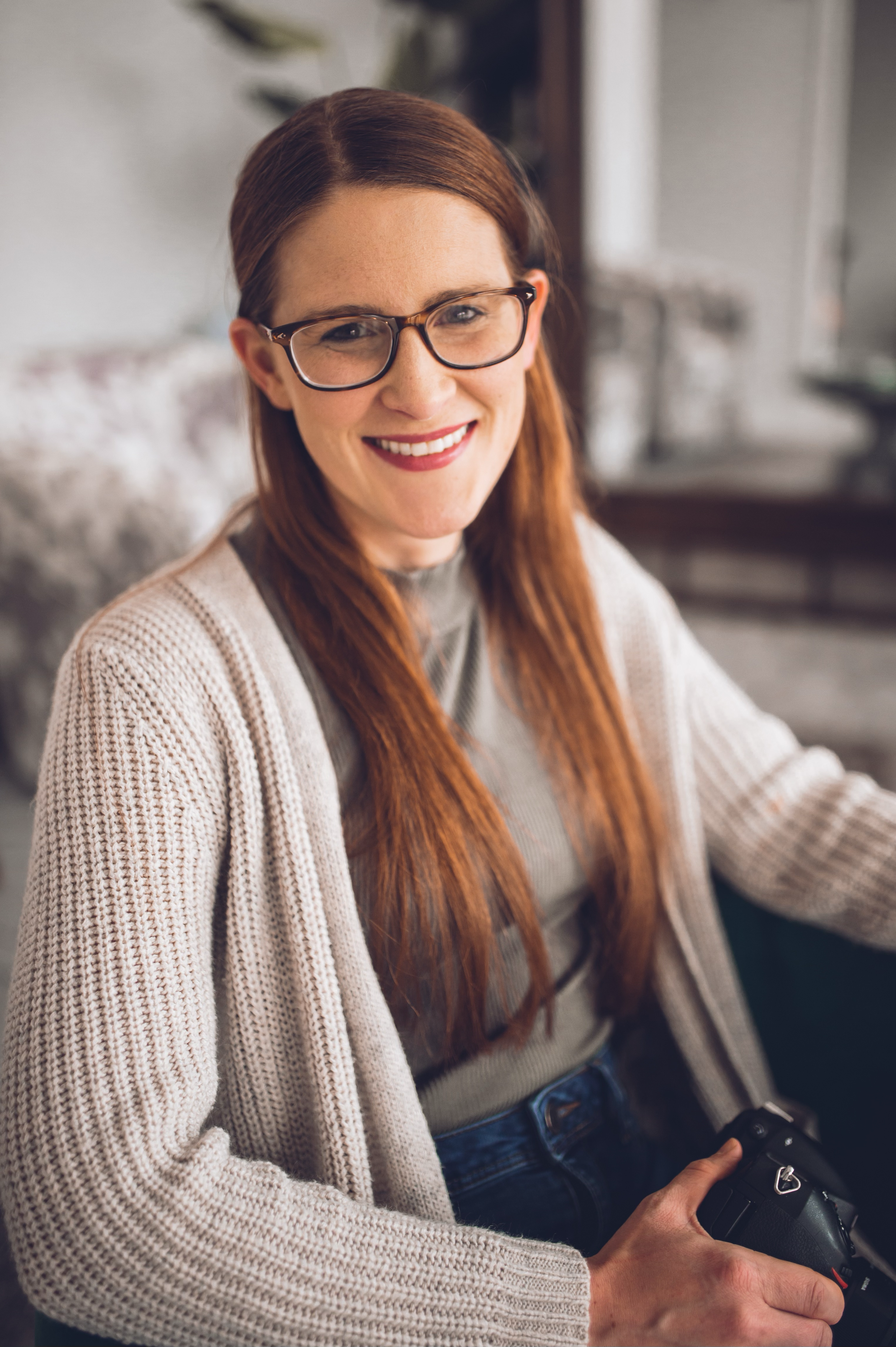 A woman with long red hair, wearing glasses, smiling, holding a camera.