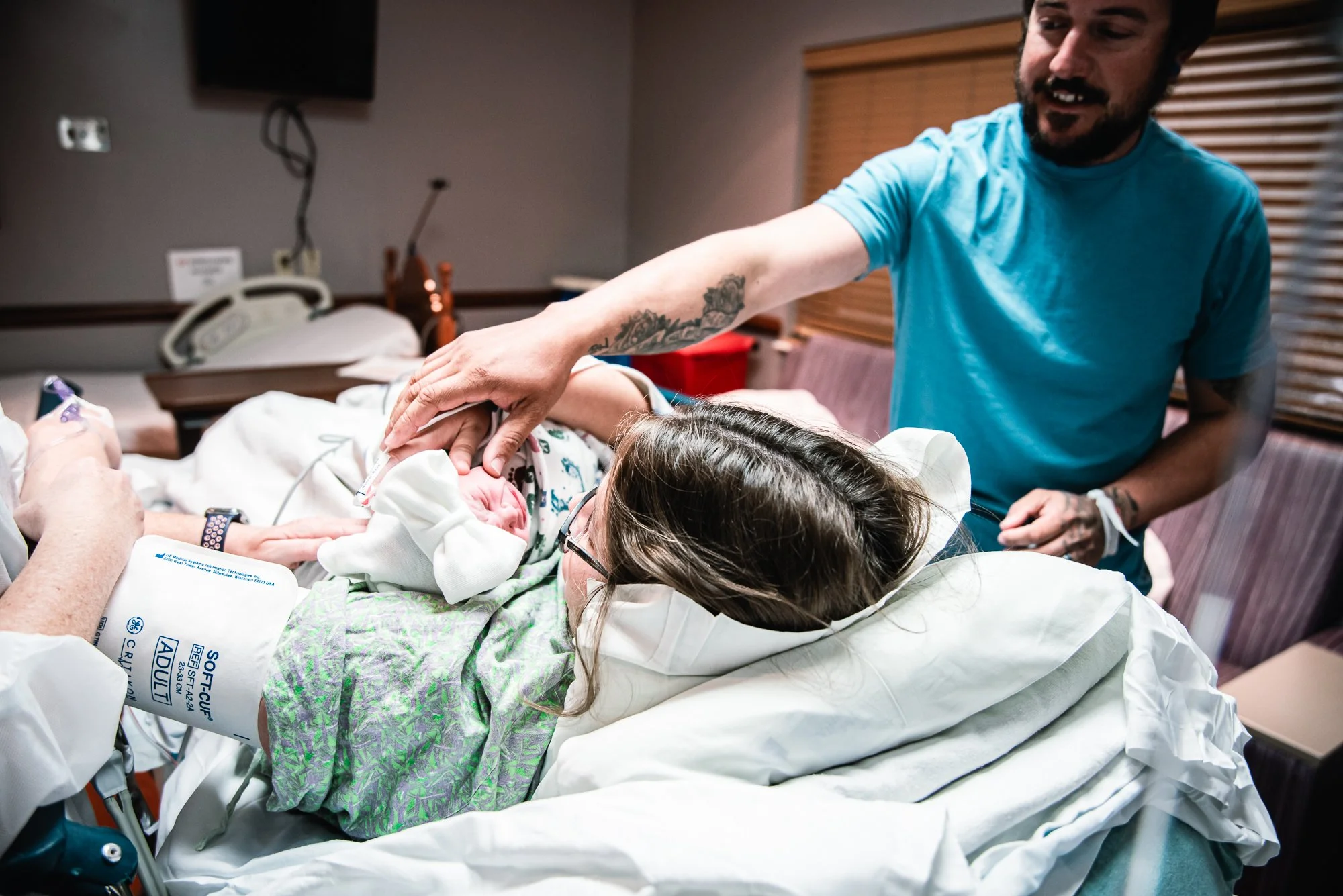 Newborn baby holding mother's finger in hospital room with doctor and visitor nearby.