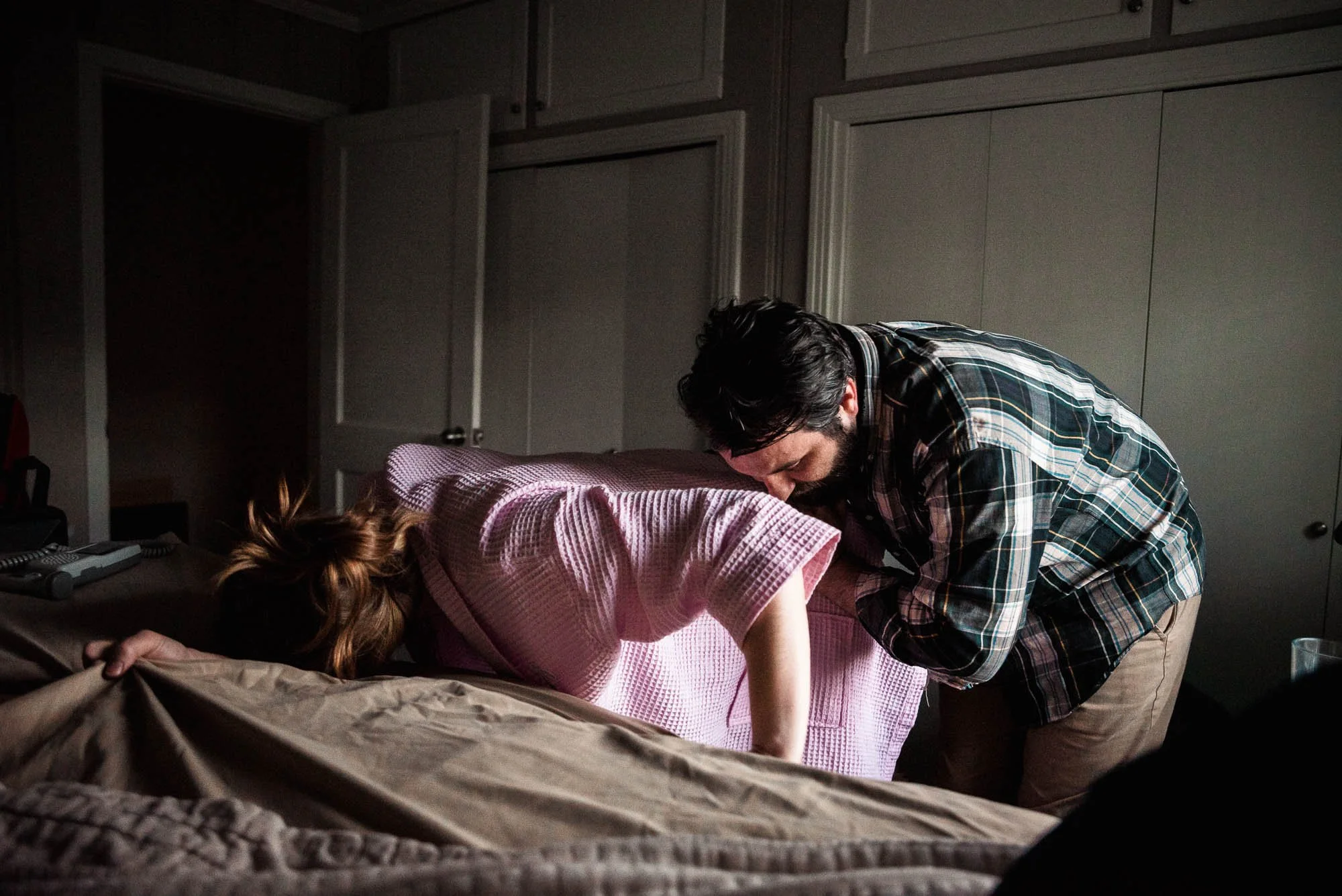 A man is performing CPR on a woman who is lying face down on a bed, with her head turned to the side in a dimly lit room.