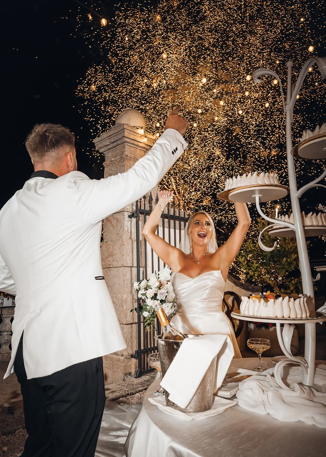 Die Braut jubelt neben dem Bräutigam und der Hochzeitstorte, während goldene Feuerwerksfunken den Nachthimmel erhellen.