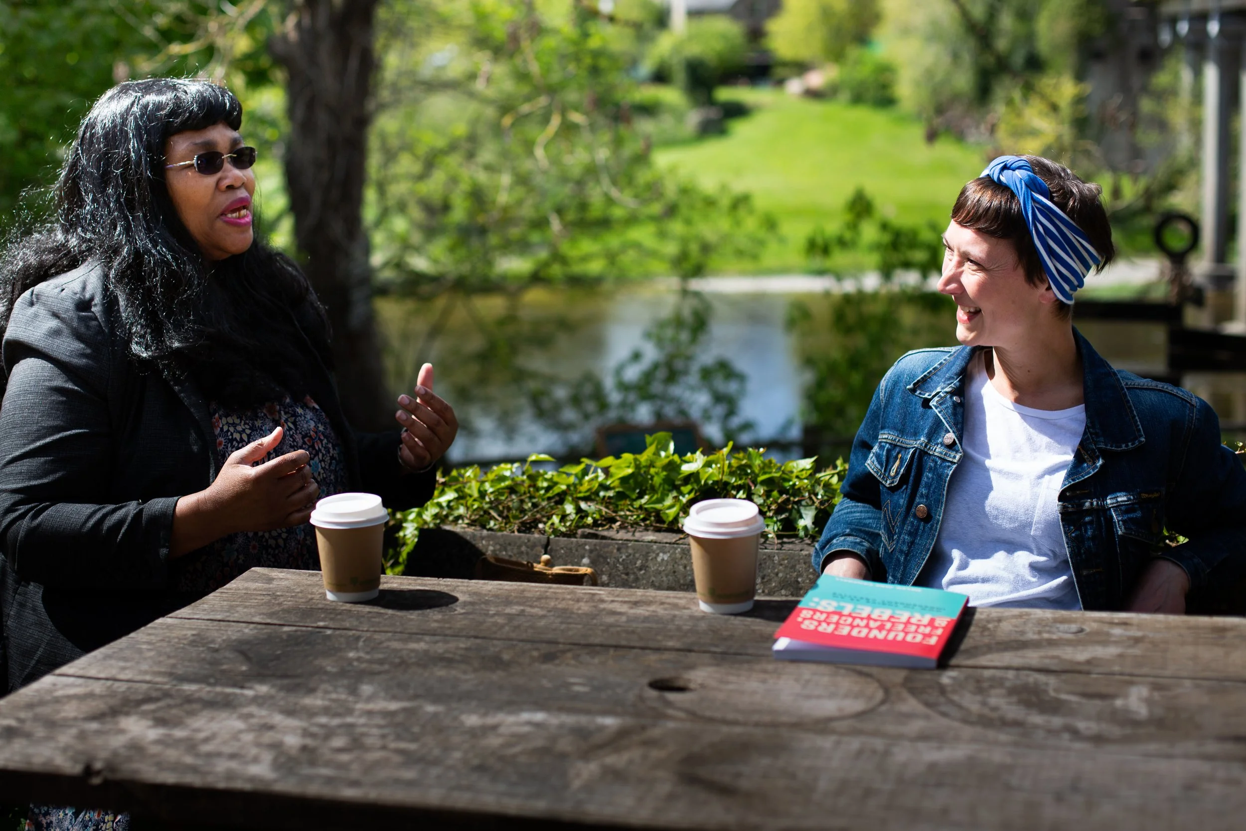 Coaching by the River Wye, a black woman with pink lipstick and long black hair with coffee, and a white woman with short brown hair and a headscarf, they are deep in conversation