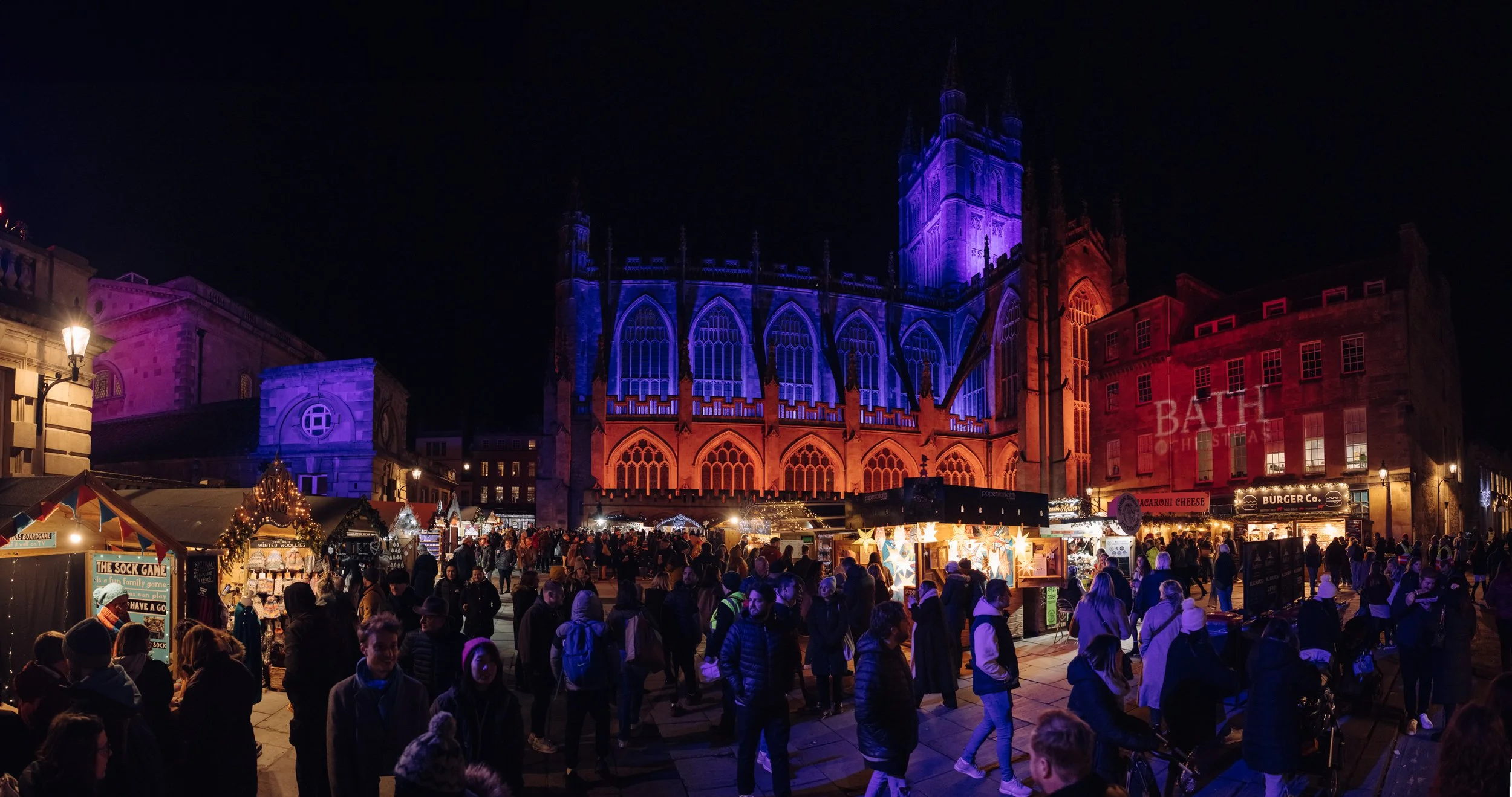 Nighttime scene of a Christmas market in Bath, England, with the Bath Abbey illuminated with colorful lights, crowded with people and market stalls.
