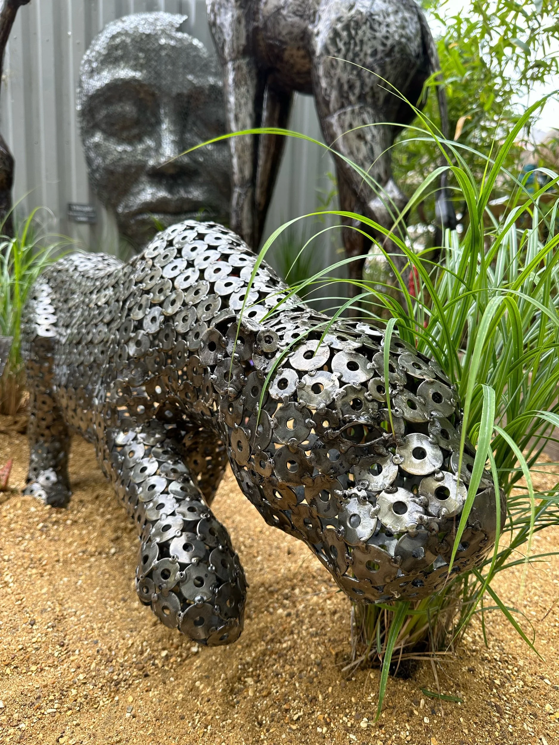 A sculpture of a dog made from metal washers and gears, surrounded by grass and sand.