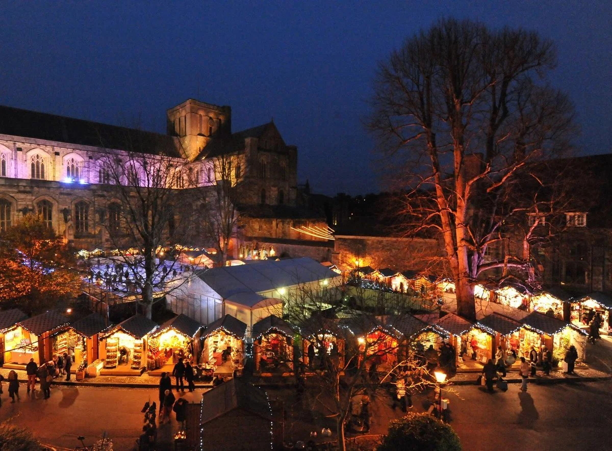 Nighttime scene of a Christmas market with numerous lit stalls, large leafless tree illuminated with lights, and a historic building in the background with colorful lights