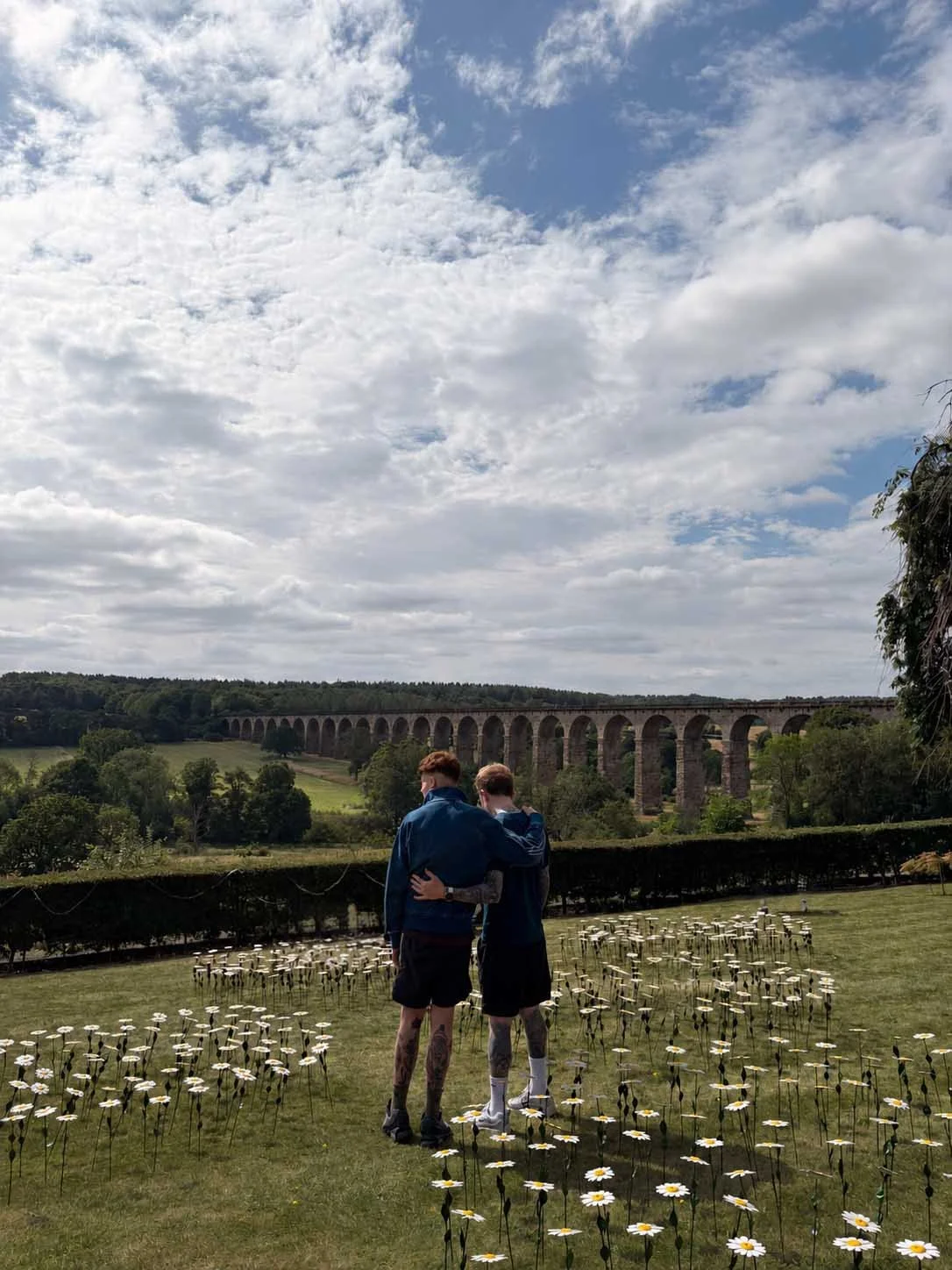 Two young men with tattoos standing close together in a field of daisies, embracing with an arm around each other's shoulders, under a partly cloudy sky, with a historic arched stone bridge in the background.