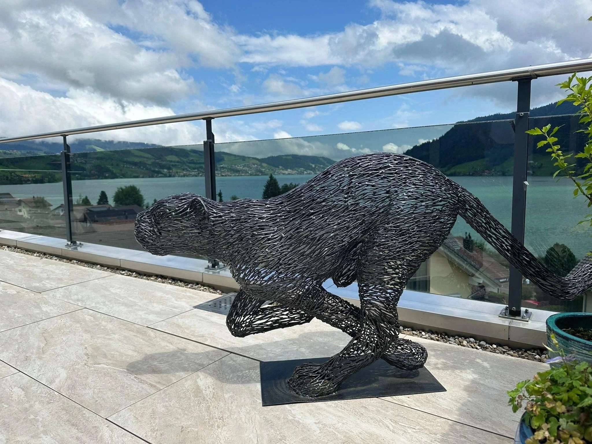 A black metal sculpture of a dog on a balcony overlooking a lake and mountains under a partly cloudy sky.