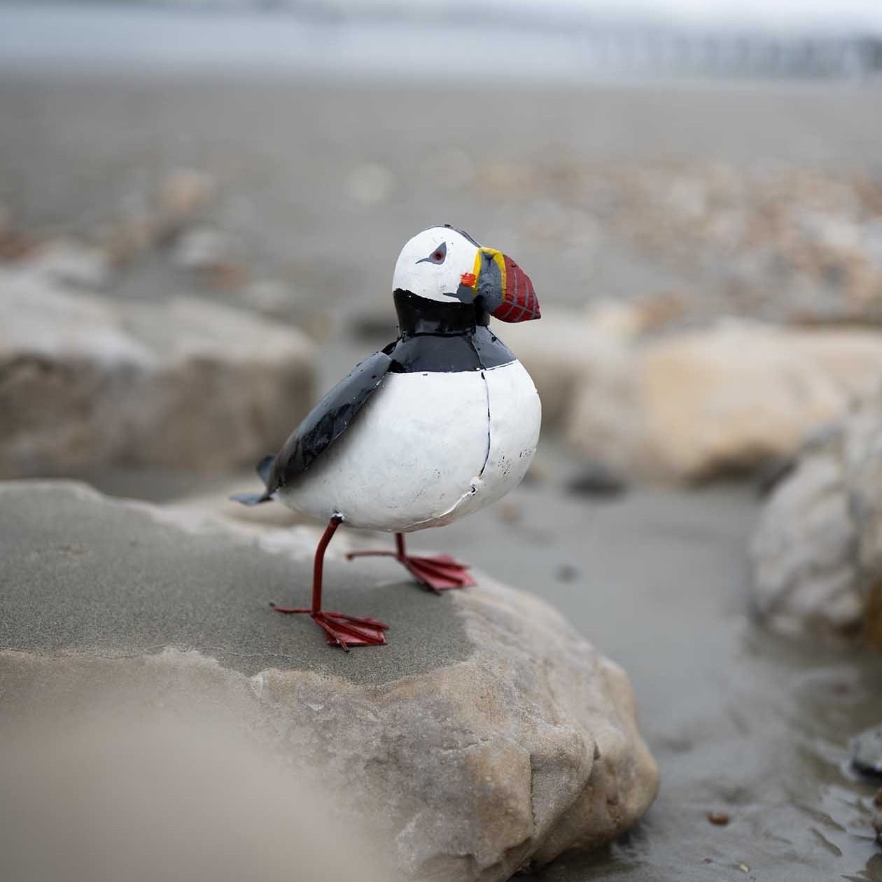 a recycled metal puffin garden ornament sat on stones on the beach with the sea in the background