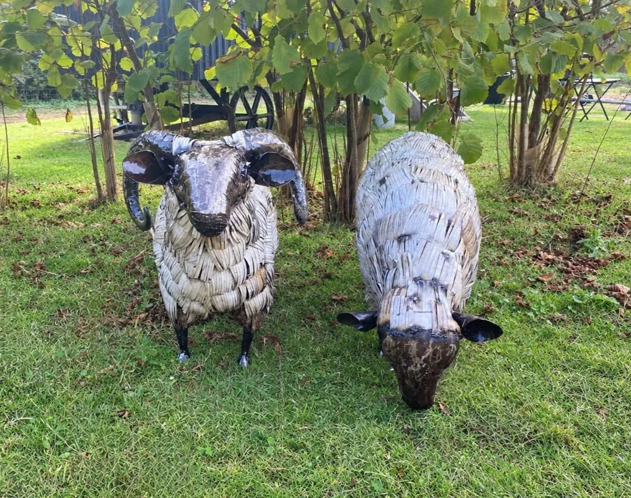Metal sculpture of a sheep and a ram in a grassy garden with trees and a wagon wheel in the background.