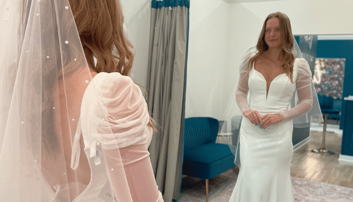 A woman in a bridal shop admires herself in the mirror, wearing a white wedding gown with sheer long sleeves and a veil. The setting is elegant and serene.