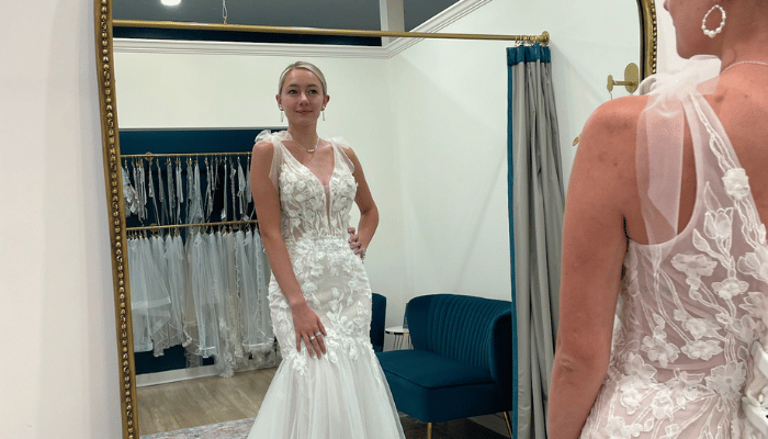 A woman in a lace wedding dress with floral details is looking in a mirror. The room is elegant, featuring a blue sofa and racks of dresses, conveying anticipation.