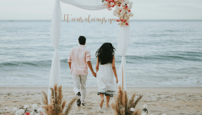 A newly wed couple holding hands walks toward the ocean under a decorated arch on a sandy beach, with white fabric and pink flowers, creating a romantic scene.