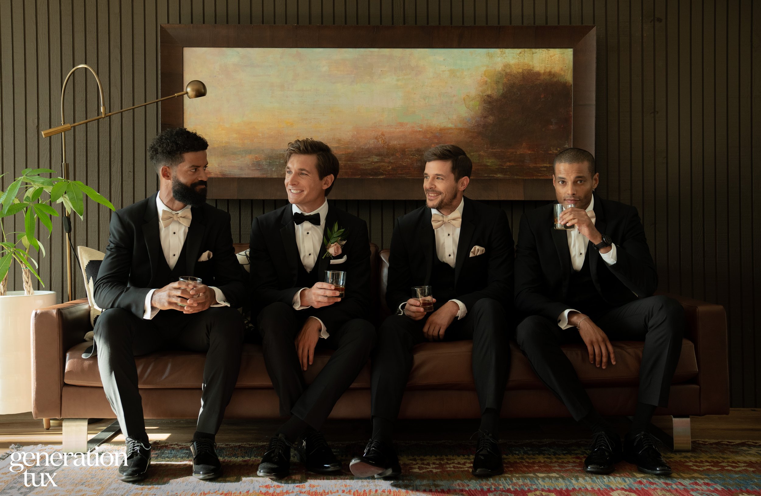 Four groomsmen in black tuxedos sitting on a couch holding drinks, showcasing formal wedding suits and modern groom style inspiration