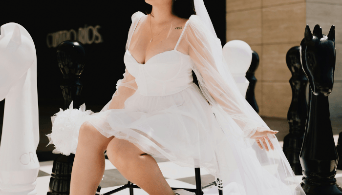 A bride in a short wedding dress, flowing white dress and veil sits on a large chessboard, surrounded by oversized black and white chess pieces. The mood is playful and elegant.