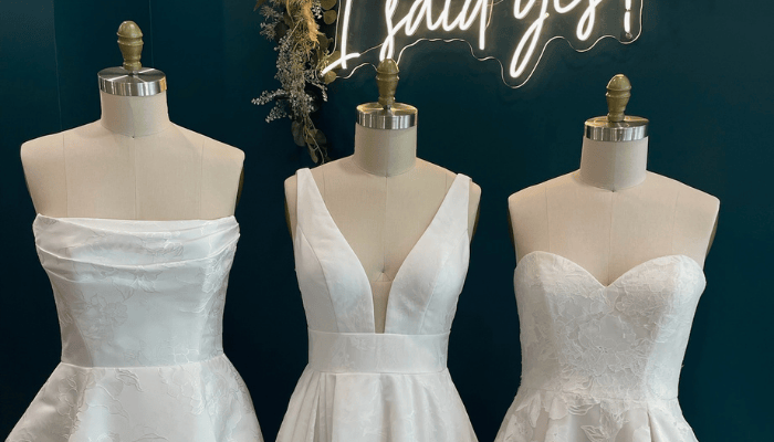 Three white wedding dresses on mannequins are displayed against a teal wall. Above them, a lit sign reads, "I said yes!" The scene feels elegant and celebratory.