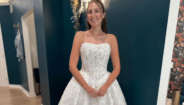 A woman joyfully models a strapless white lace wedding gown with intricate floral patterns, set in a chic bridal boutique against dark green walls.