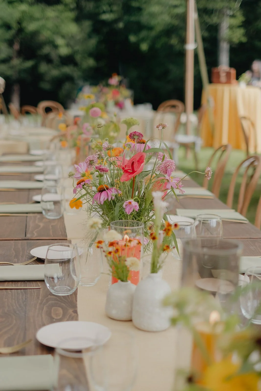 A long wooden table set outdoors with plates, glasses, and napkins boasts colorful wildflower arrangements created by a Hudson Valley wedding florist. Wooden chairs surround it, while the background showcases a grassy area with trees and a table.
