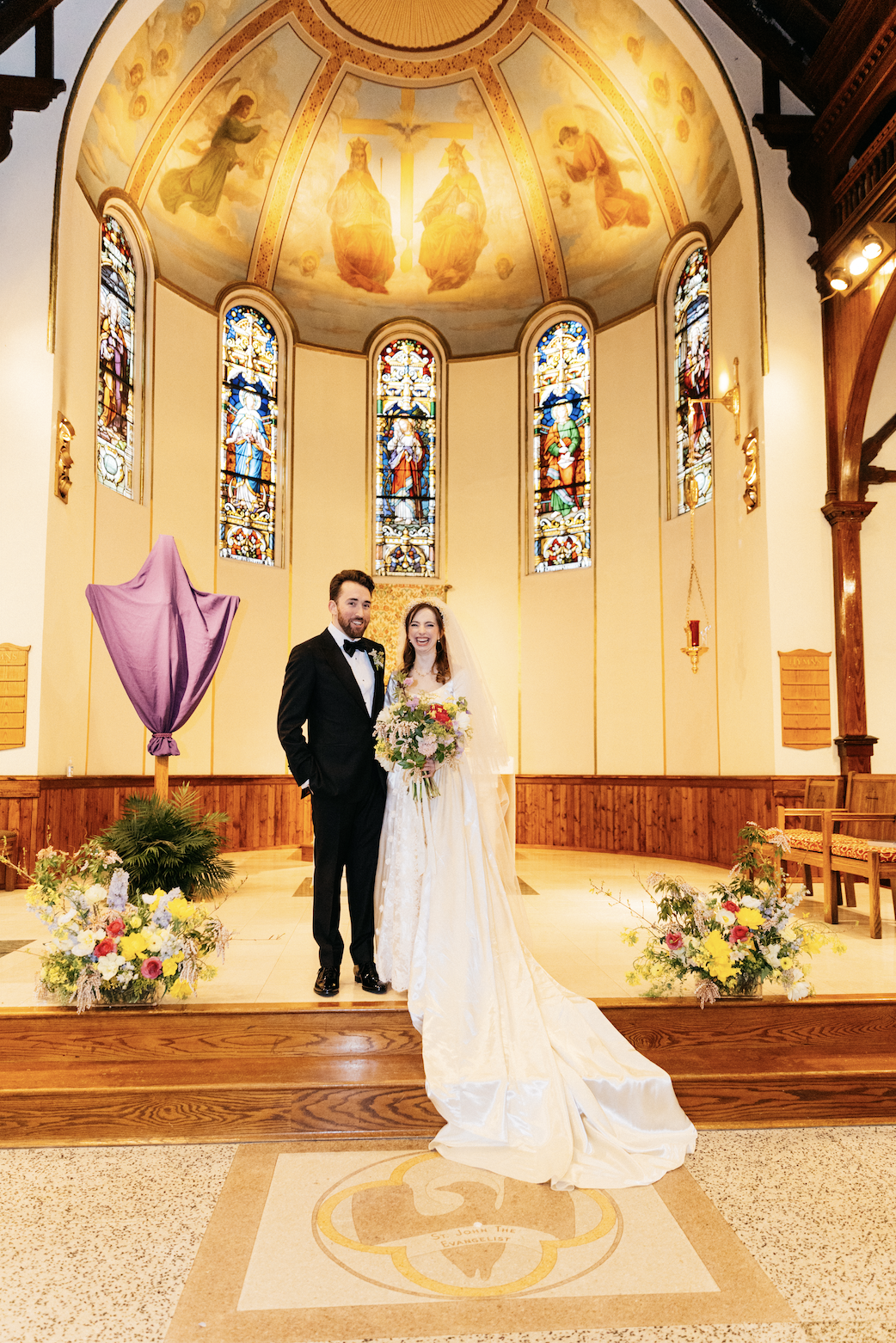 Bride and groom standing inside a light-filled church while holding bold bridal bouquet and two asymmetrical, arrangements on either side of them.