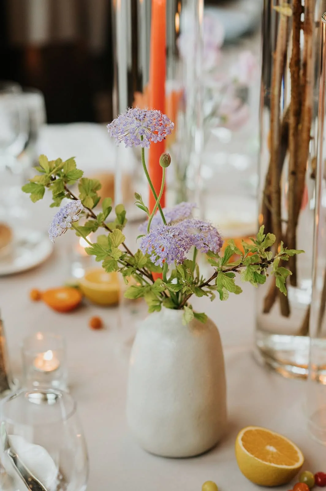 Ceramic bud vase with play purple blossoms and robust leaves sits on wedding table surrounded by candles and fruit accents 