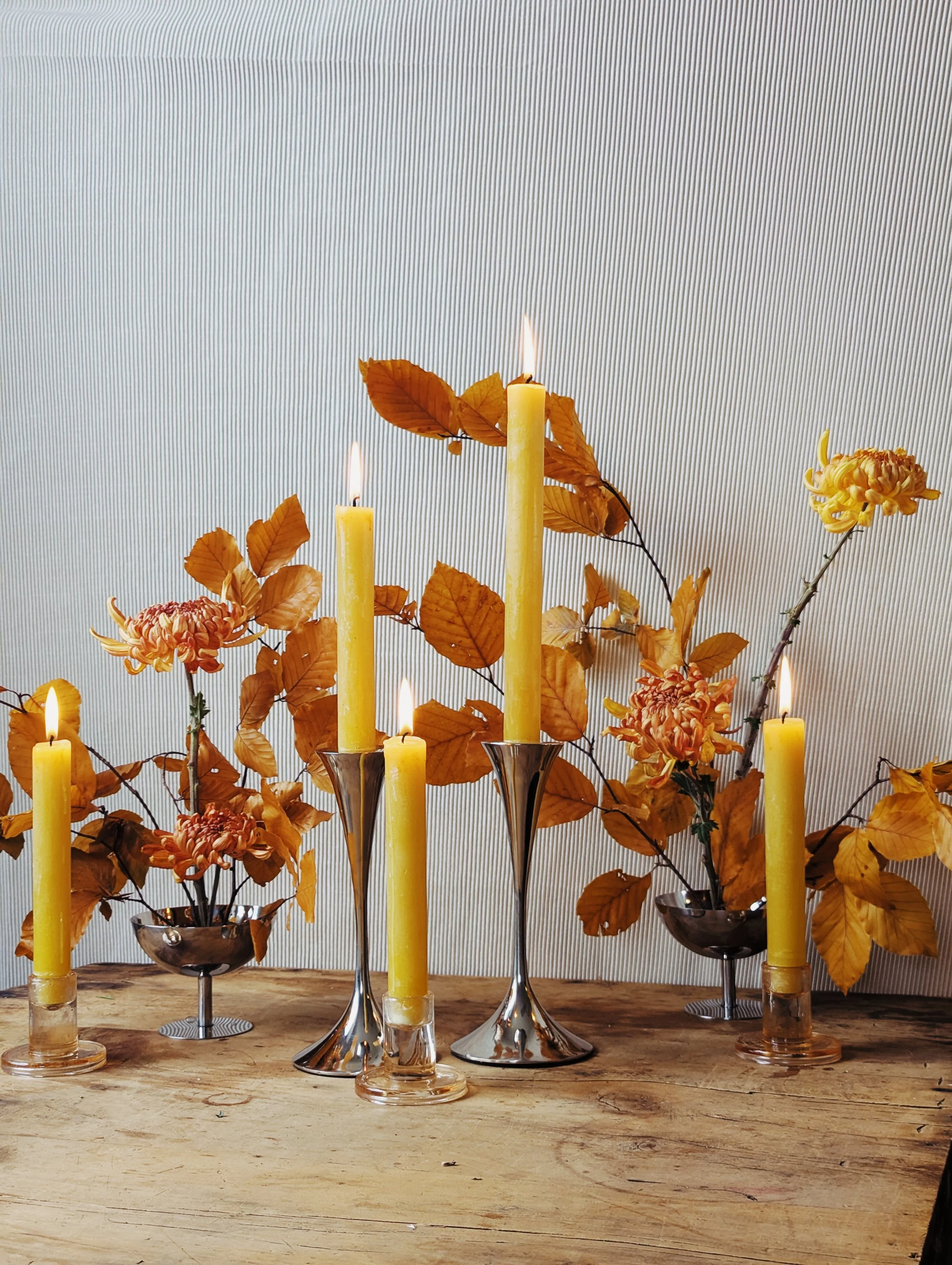   Elegant yellow taper candles on silver and glass holders in front of delicate orange foliage composition in metallic cups 